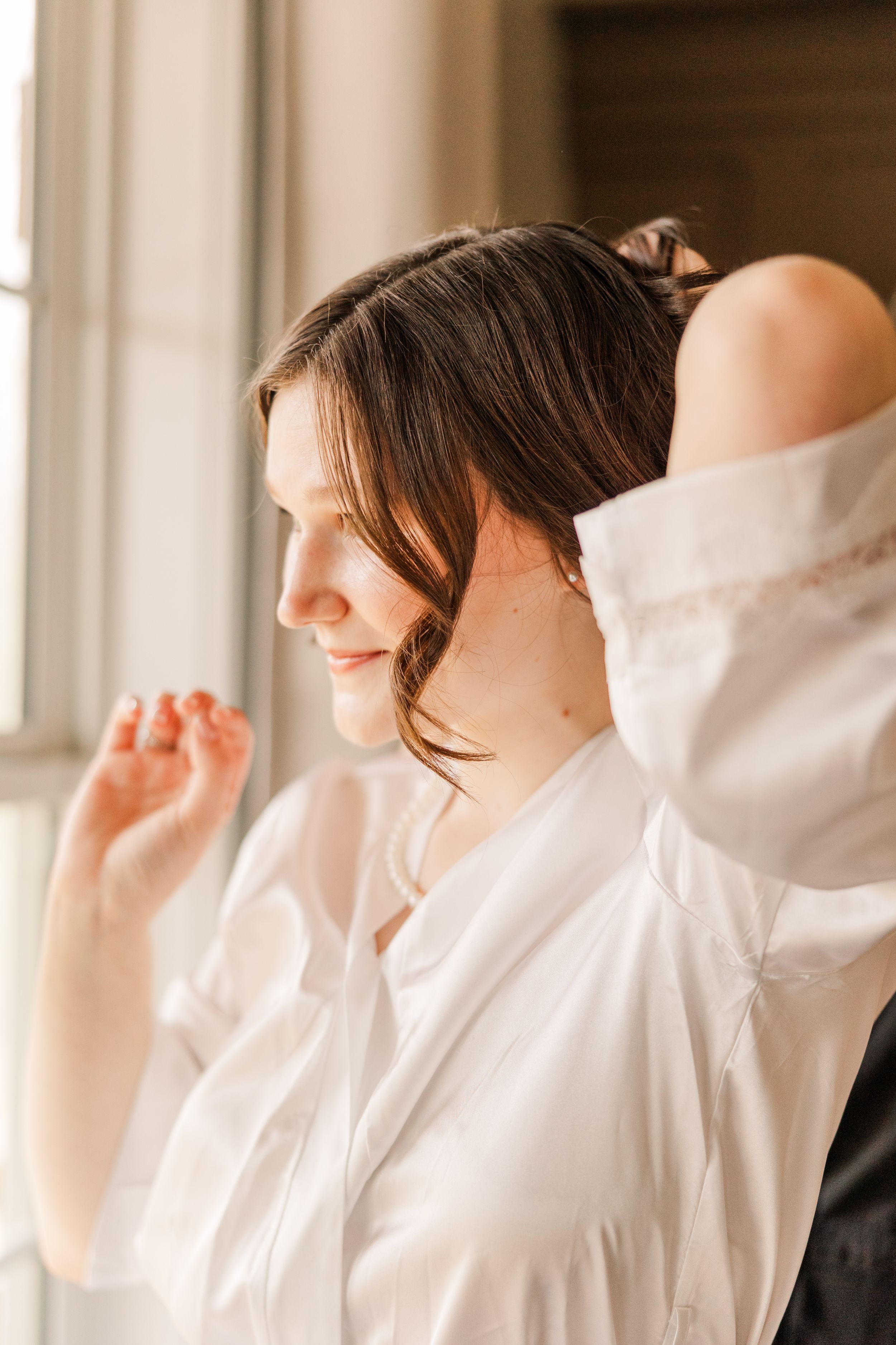 Bride in white robe gazes out window during wedding day preparations.