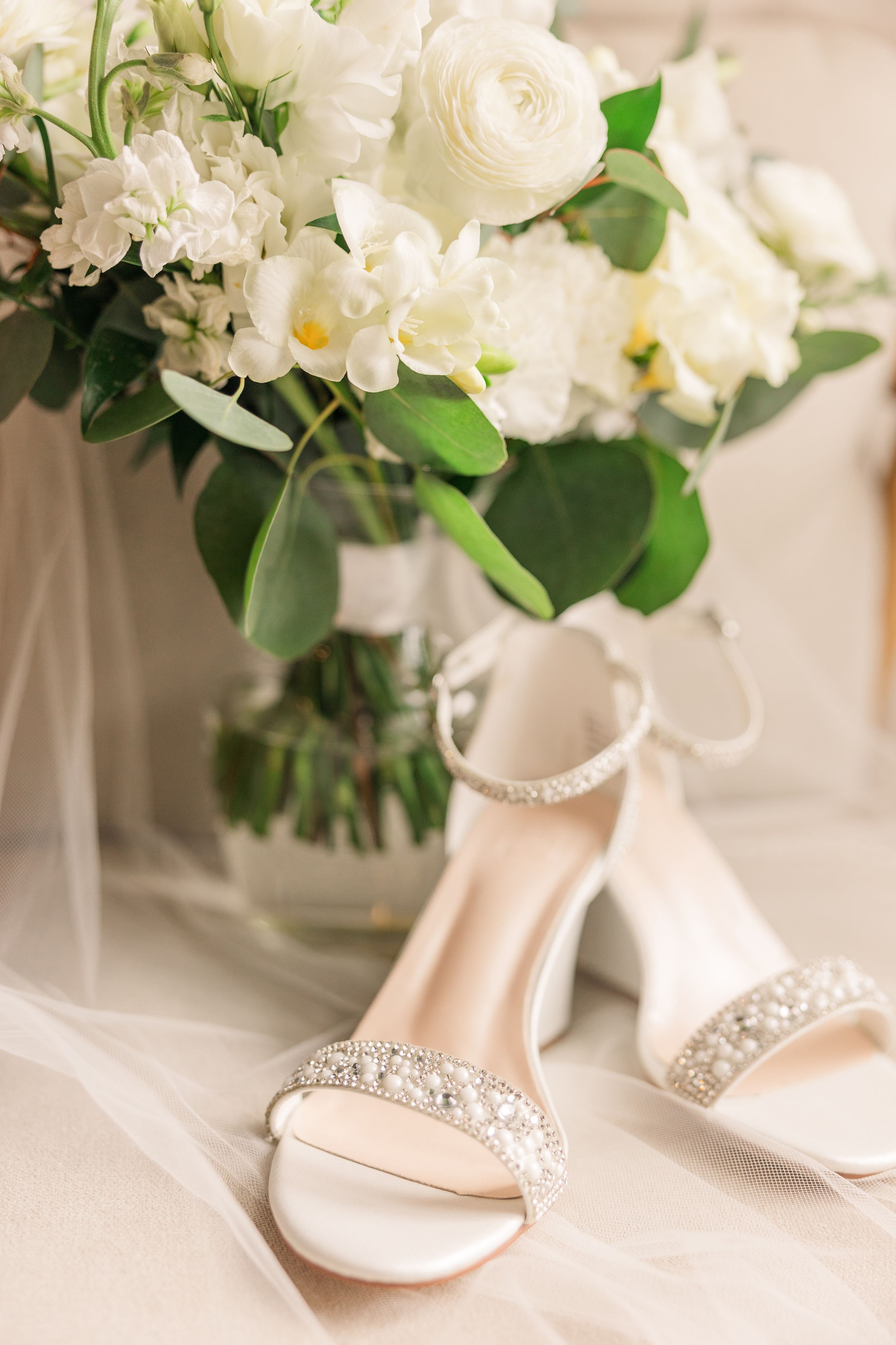 Elegant bridal bouquet of white roses and flowers next to sparkly wedding heels on white tulle fabric.