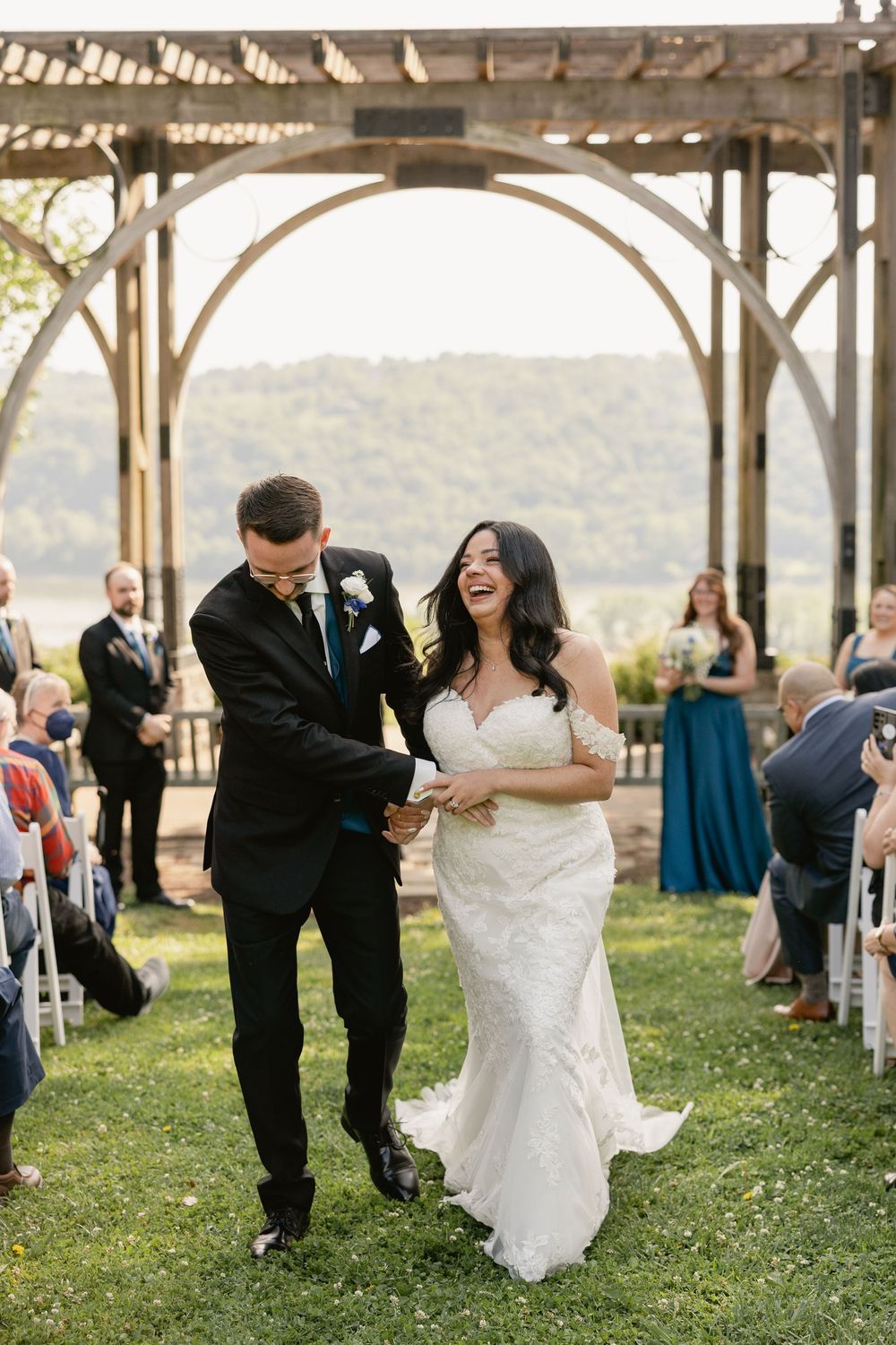Joyful bride and groom walk down outdoor wedding aisle under rustic wooden pergola with scenic river views.