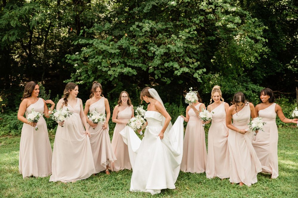 Bridesmaids in blush pink dresses walk together on grass during an elegant outdoor wedding photo session.