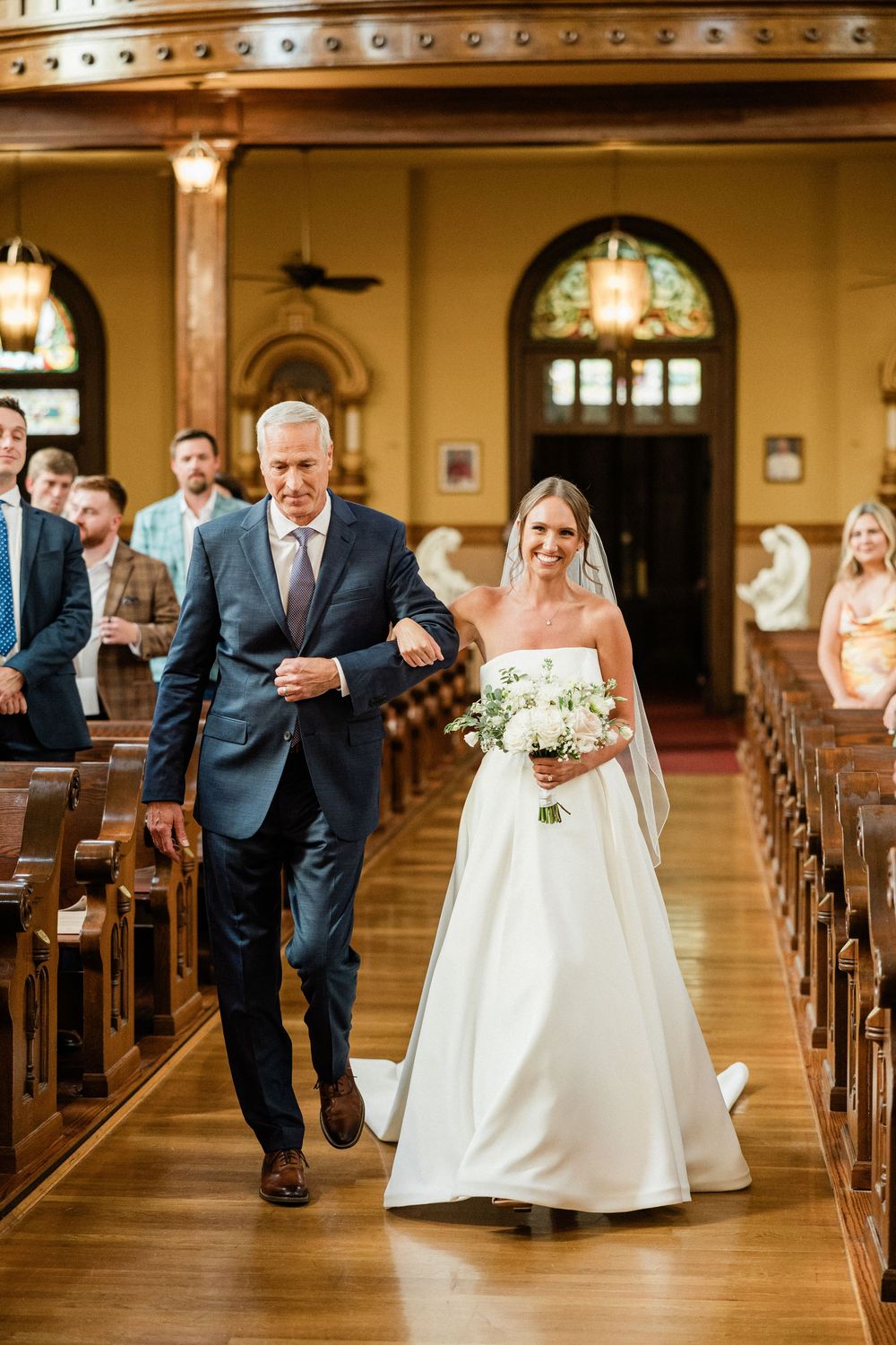 A wedding party playfully runs down the aisle of a traditional church with wooden pews and warm lighting.