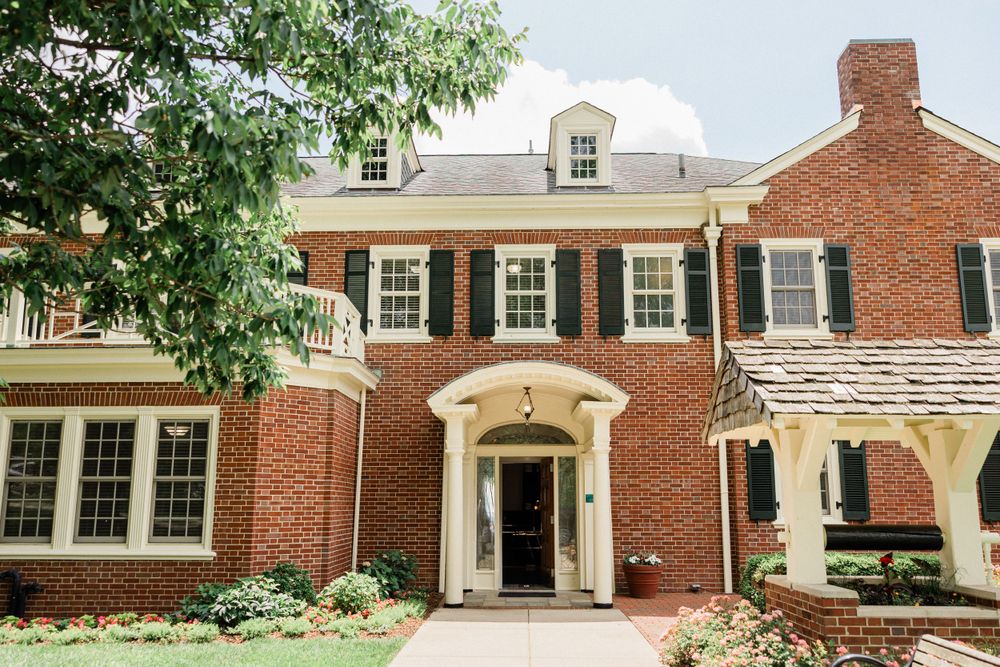 Elegant brick mansion with black shutters and a grand white entrance portico surrounded by landscaped gardens.