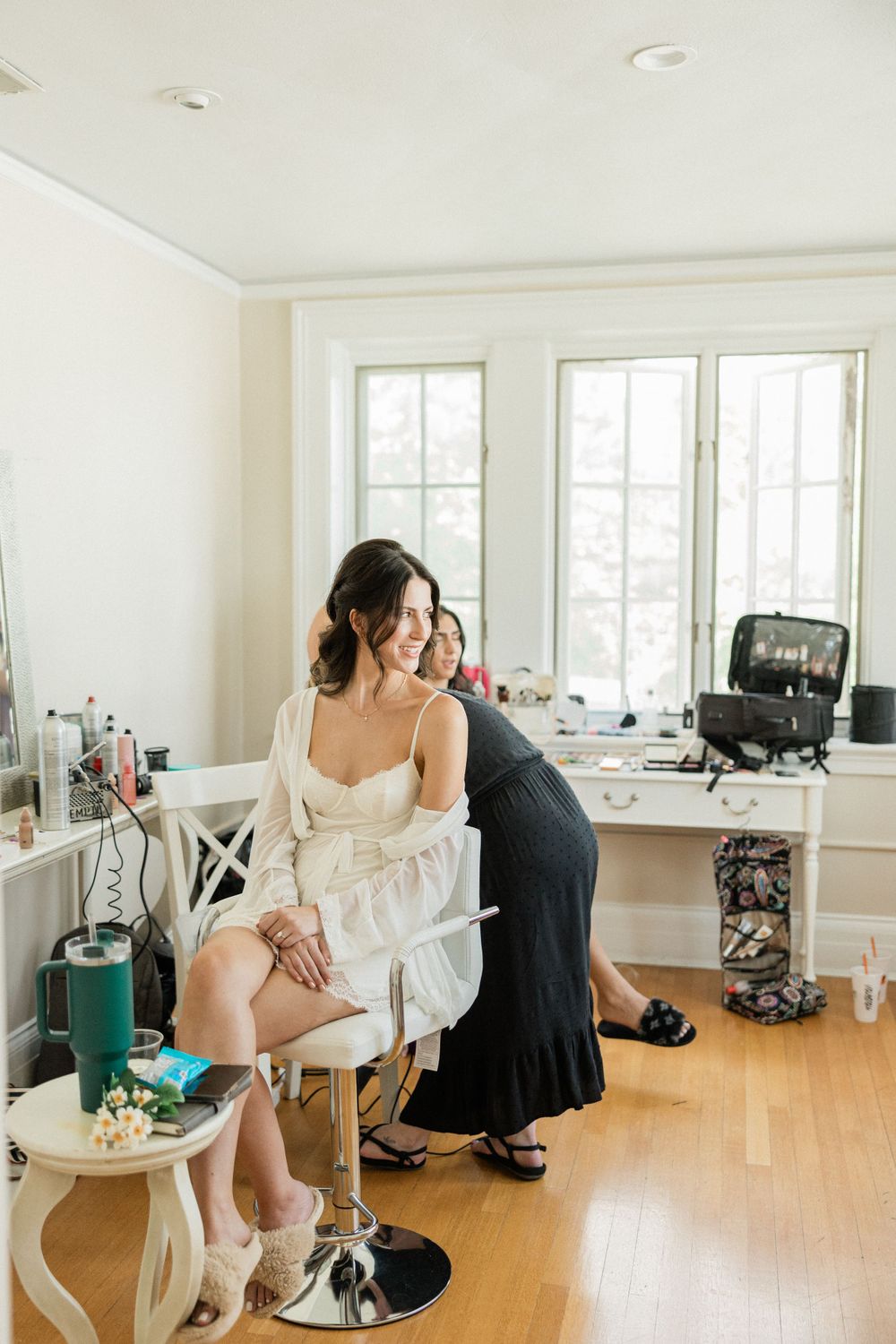 Bright sunlit room with white walls and hardwood floors being used as a bridal preparation space.
