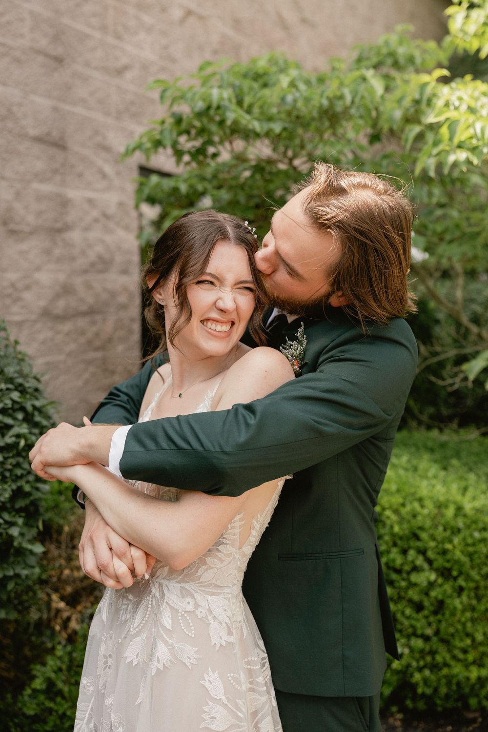 Groom in green suit hugging and kissing bride in lace wedding dress, both laughing outdoors.