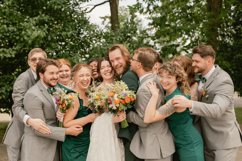 Wedding party in green dresses and gray suits surrounds smiling bride holding colorful bouquet outdoors.