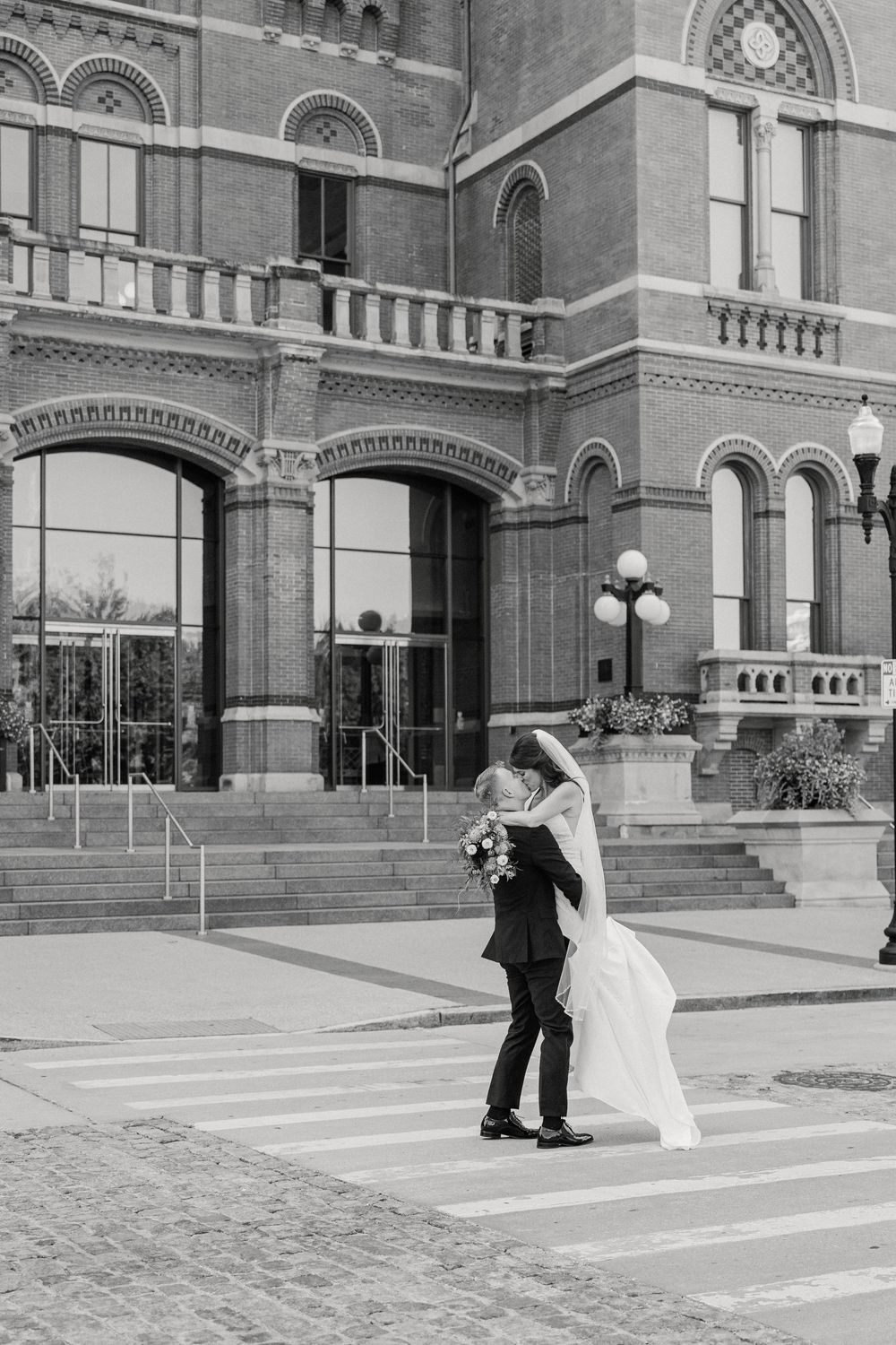 Black and white photo of a bride and groom sharing a romantic kiss outside a grand brick building on their wedding day.