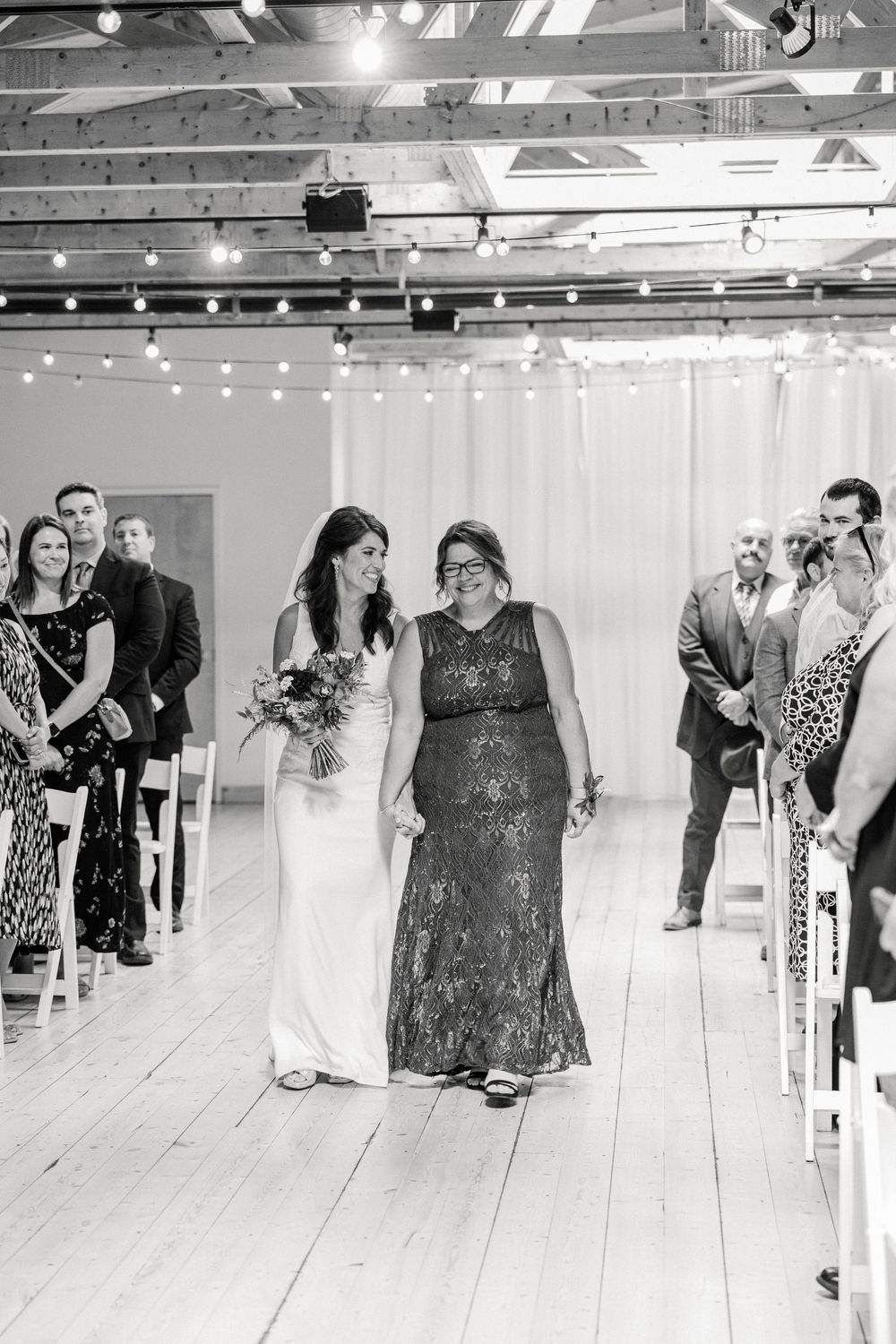 Bride walks down the aisle with a guest at an indoor wedding ceremony with string lights.