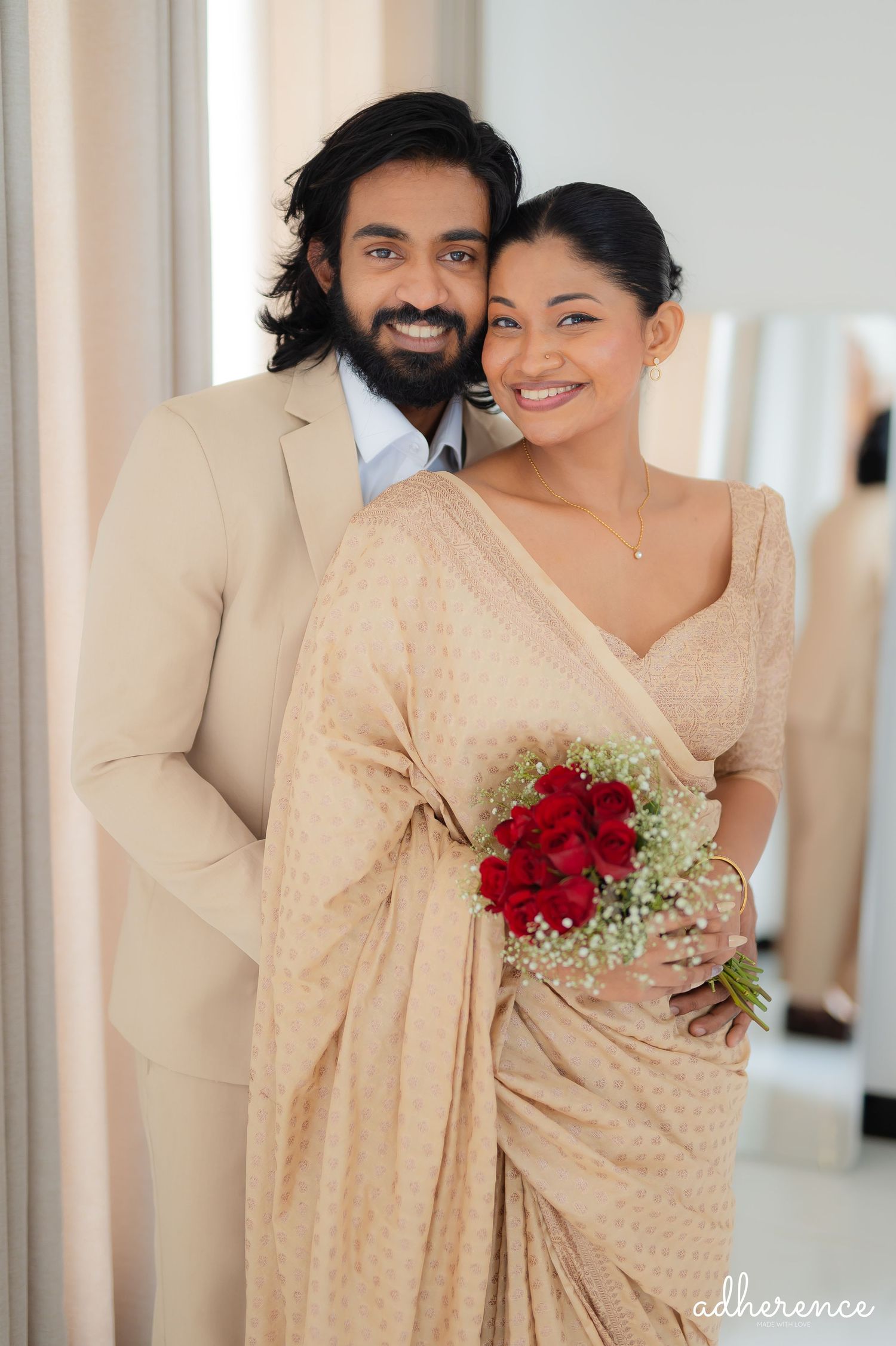 Couple in traditional beige attire pose with red bouquet in bright interior setting.