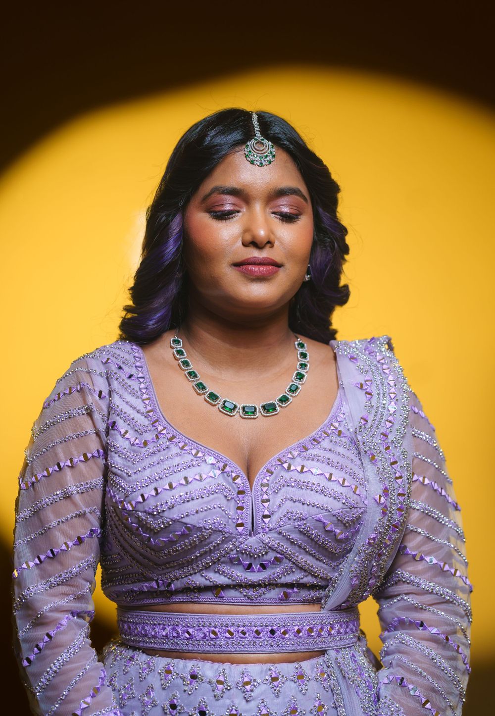 Woman in lavender beaded lehenga poses against yellow backdrop wearing traditional jewelry.