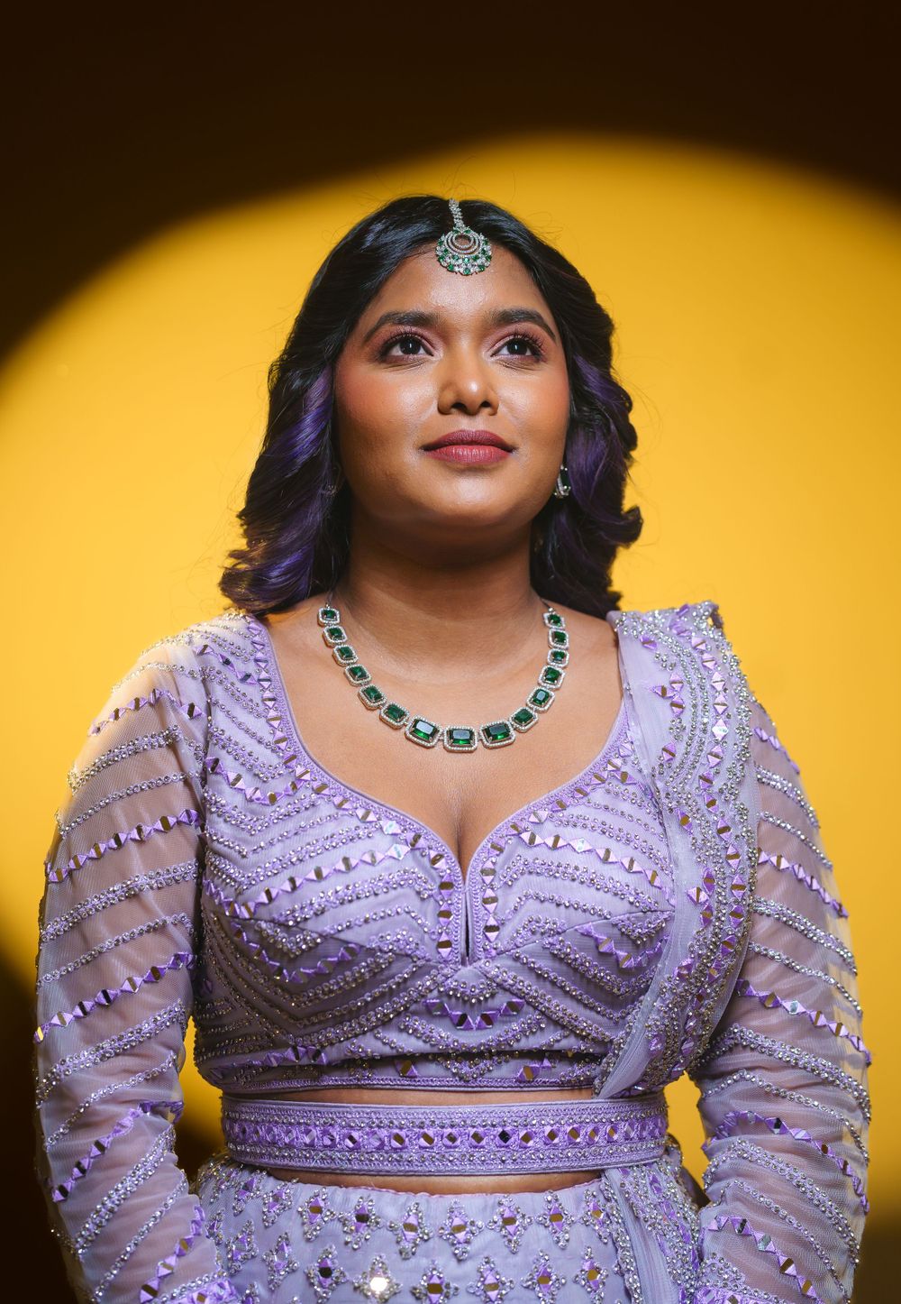 Woman in ornate purple lehenga with beaded details poses against dramatic yellow background.