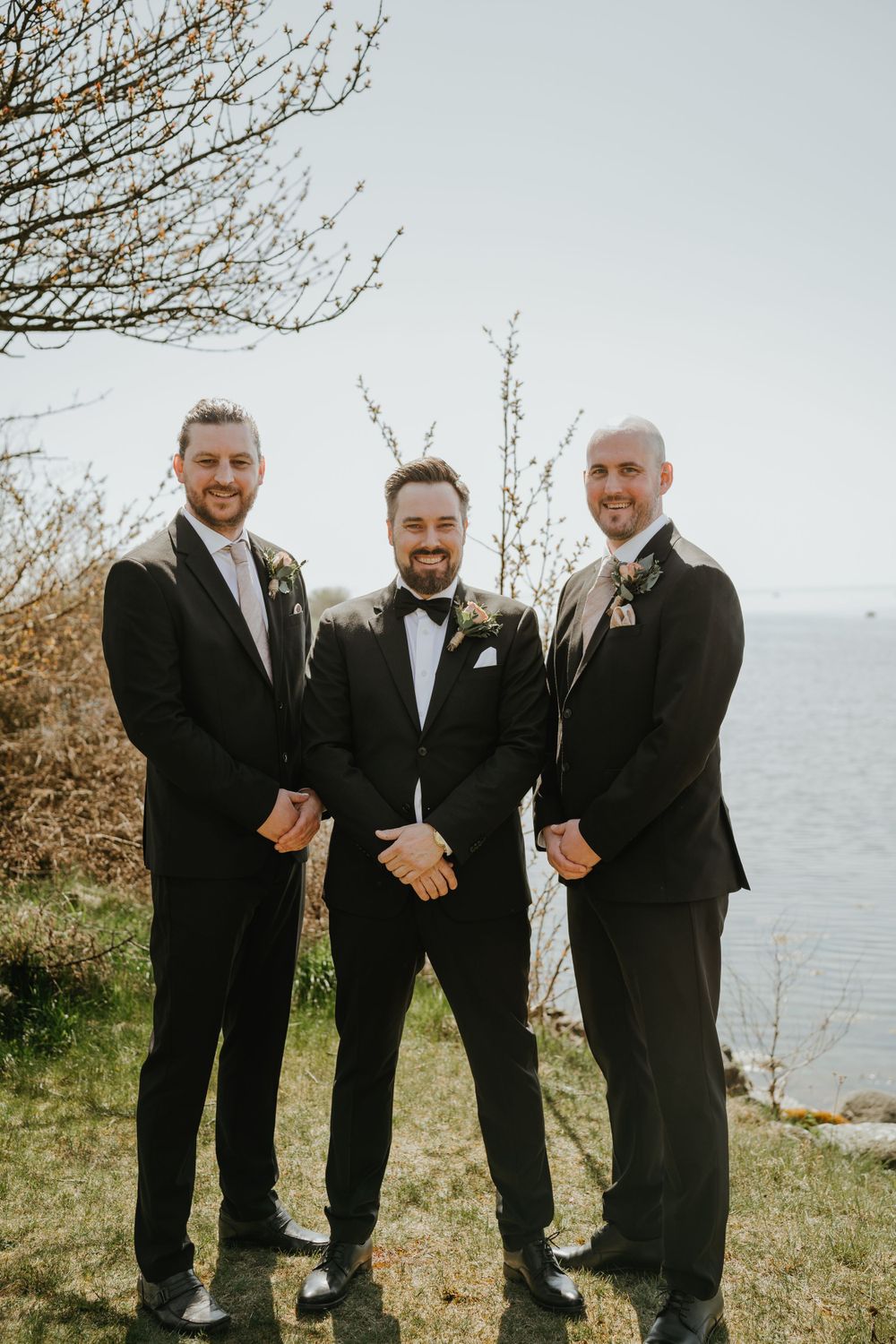 Three groomsmen in dark suits pose together on a coastal hillside during a sunny wedding day.