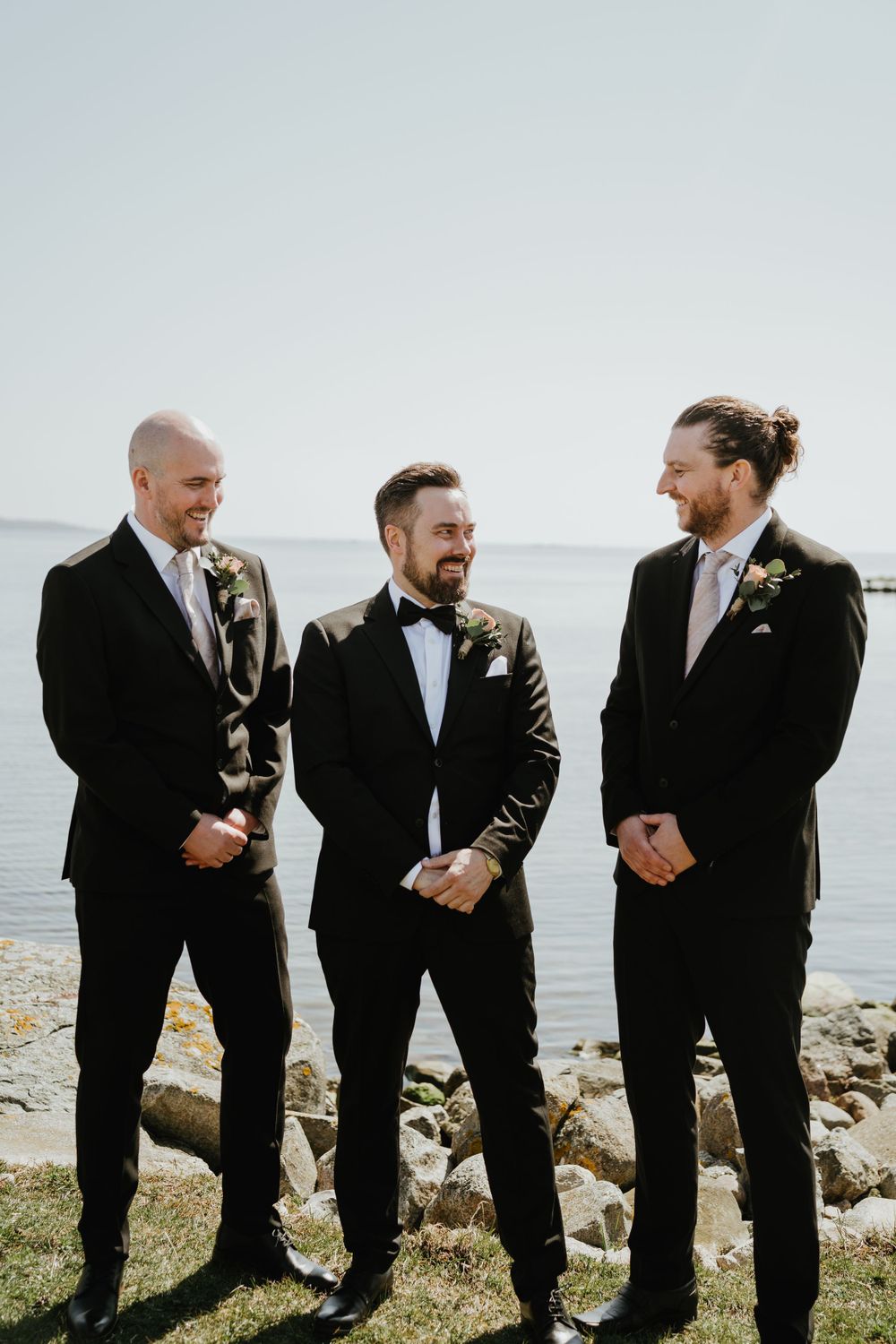 Three groomsmen share a candid moment while standing on rocky coastline in formal attire.