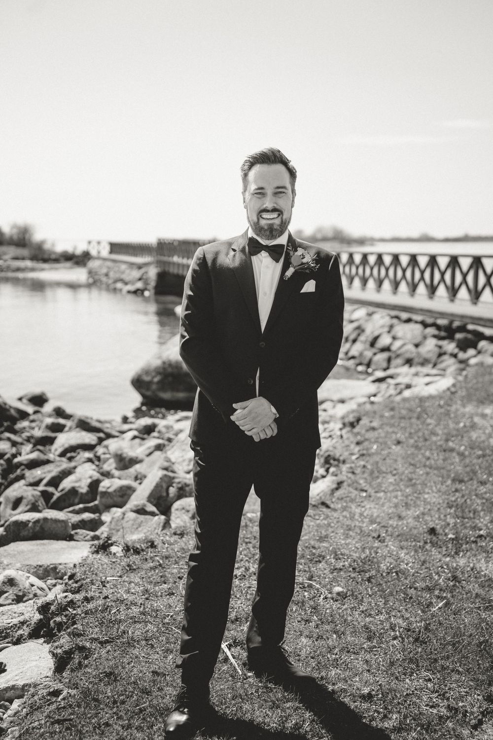 Classic black and white portrait of groom in tuxedo standing by coastal rocks.