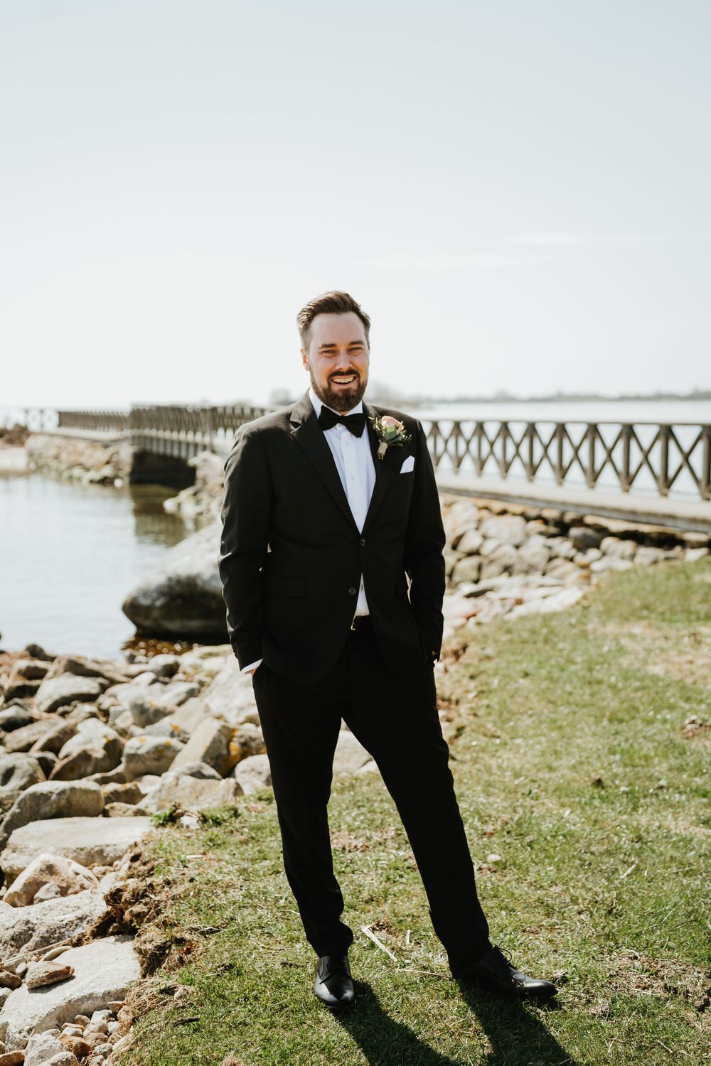 Groom in classic tuxedo poses near wooden bridge on rocky coastline.