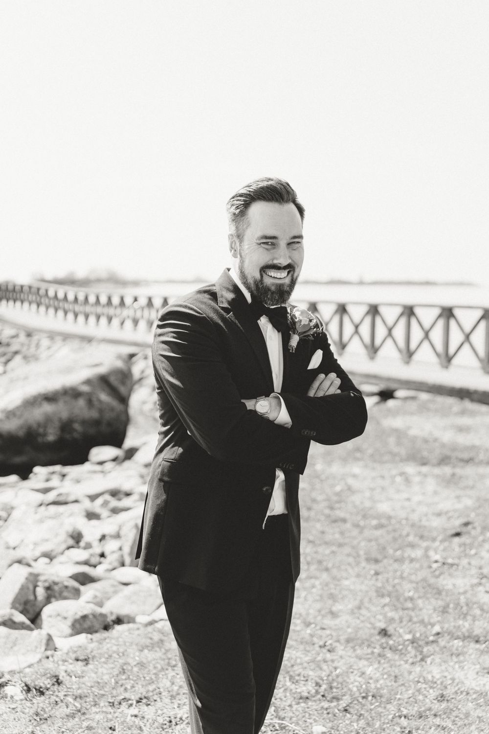 Black and white portrait of groom smiling with arms crossed near seaside bridge.