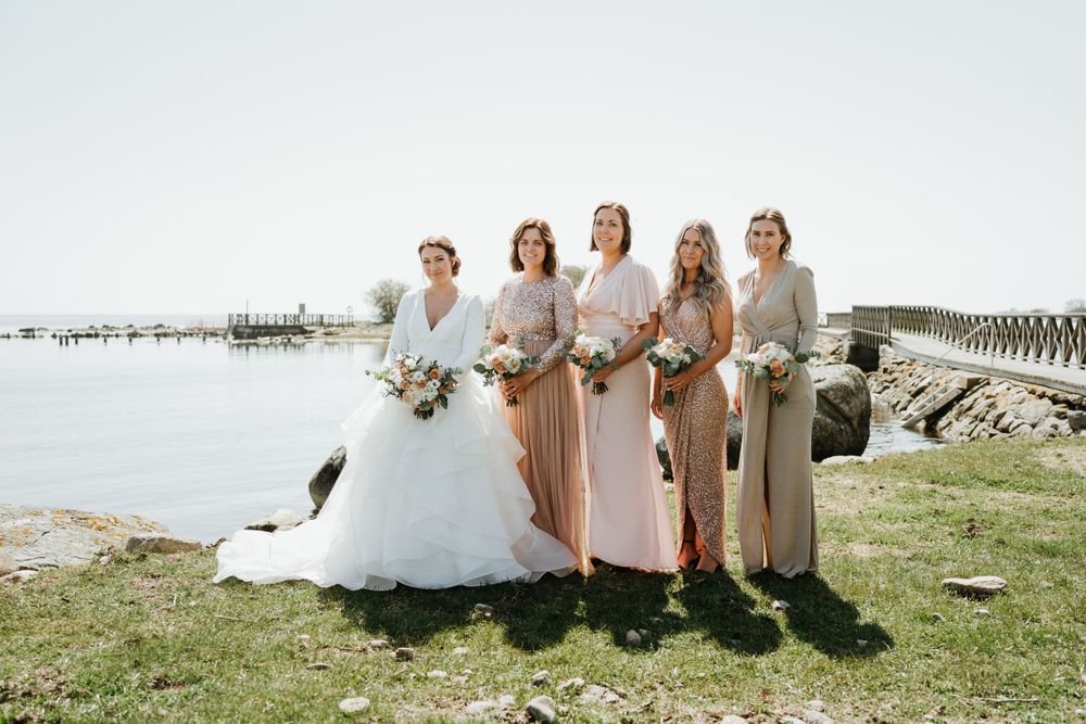 Bride and bridesmaids in pastel dresses pose together by the ocean on sunny wedding day.