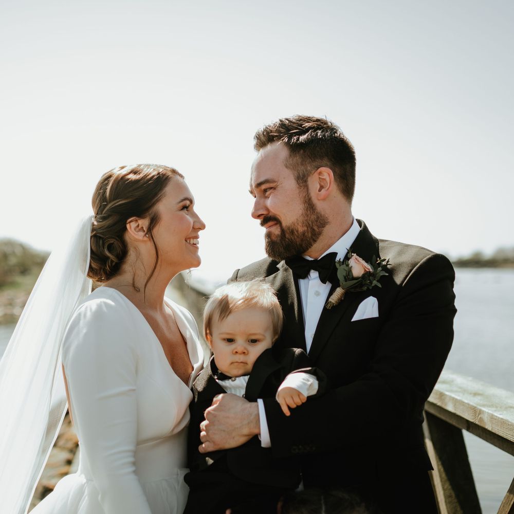 Wedding couple and small child pose together on wooden dock by water.