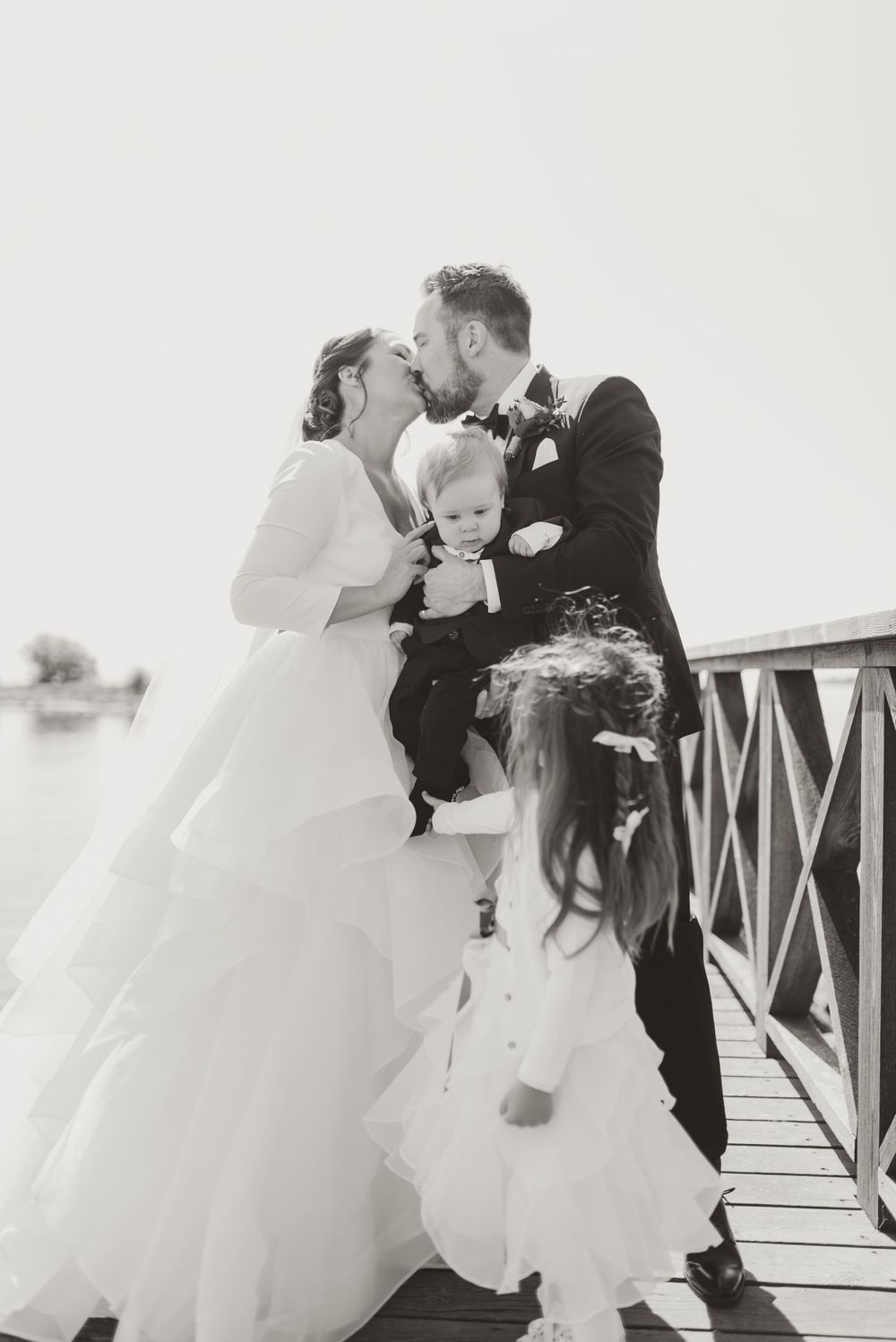 Intimate black and white wedding moment with couple and children on wooden bridge.