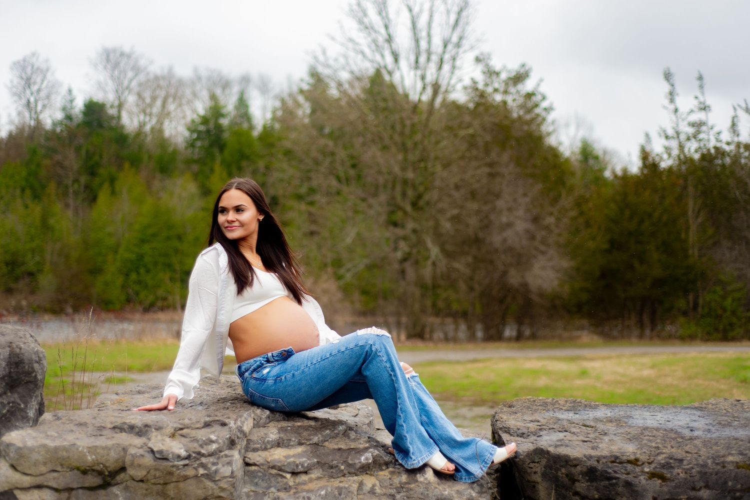 Maternity photoshoot of woman in white top and jeans sitting on large rock formation.