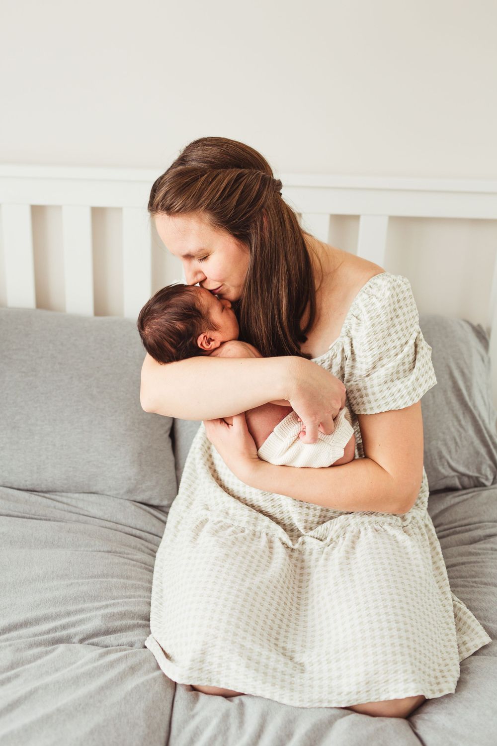 Mother in gingham dress holding sleeping newborn on grey bed.