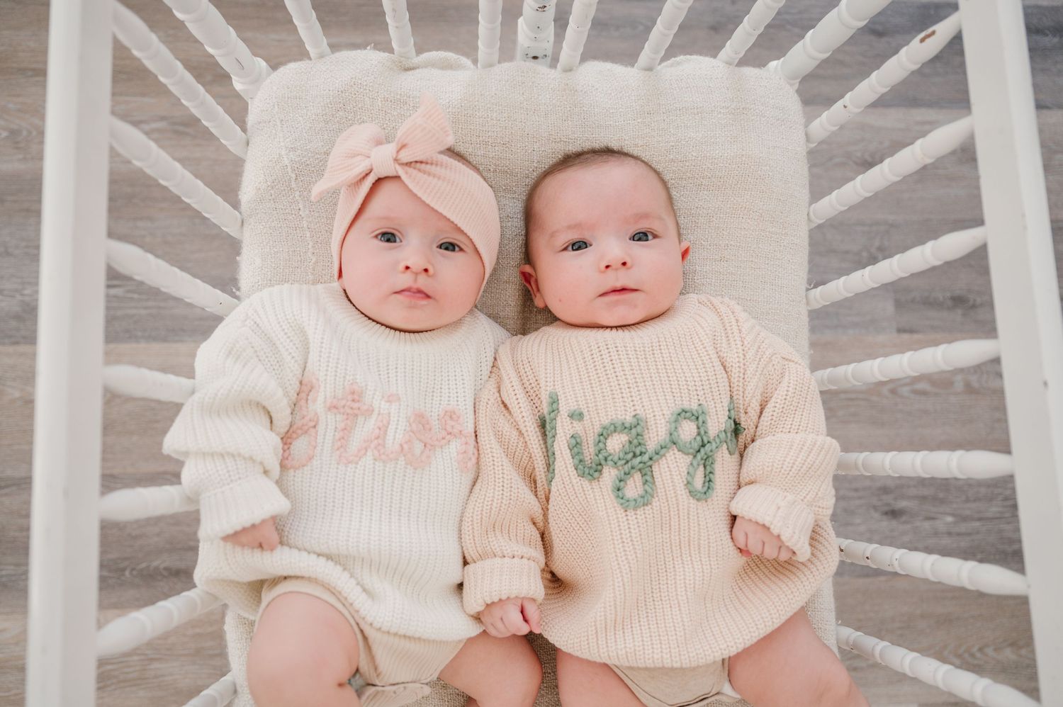 Two babies lying side by side in a crib wearing matching monogrammed sweaters.