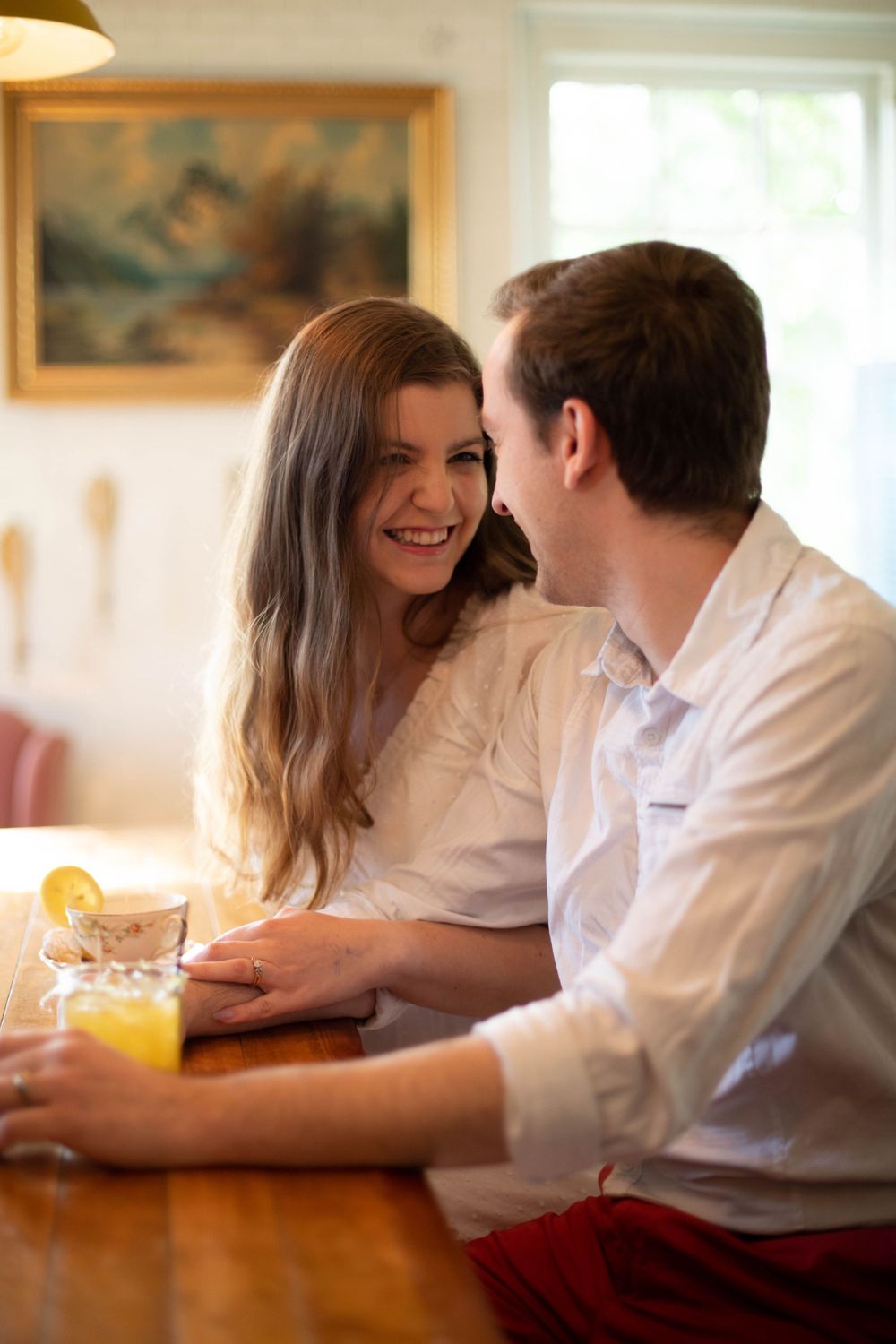 Casual couple sharing laughs at rustic wooden table in sunlit room.