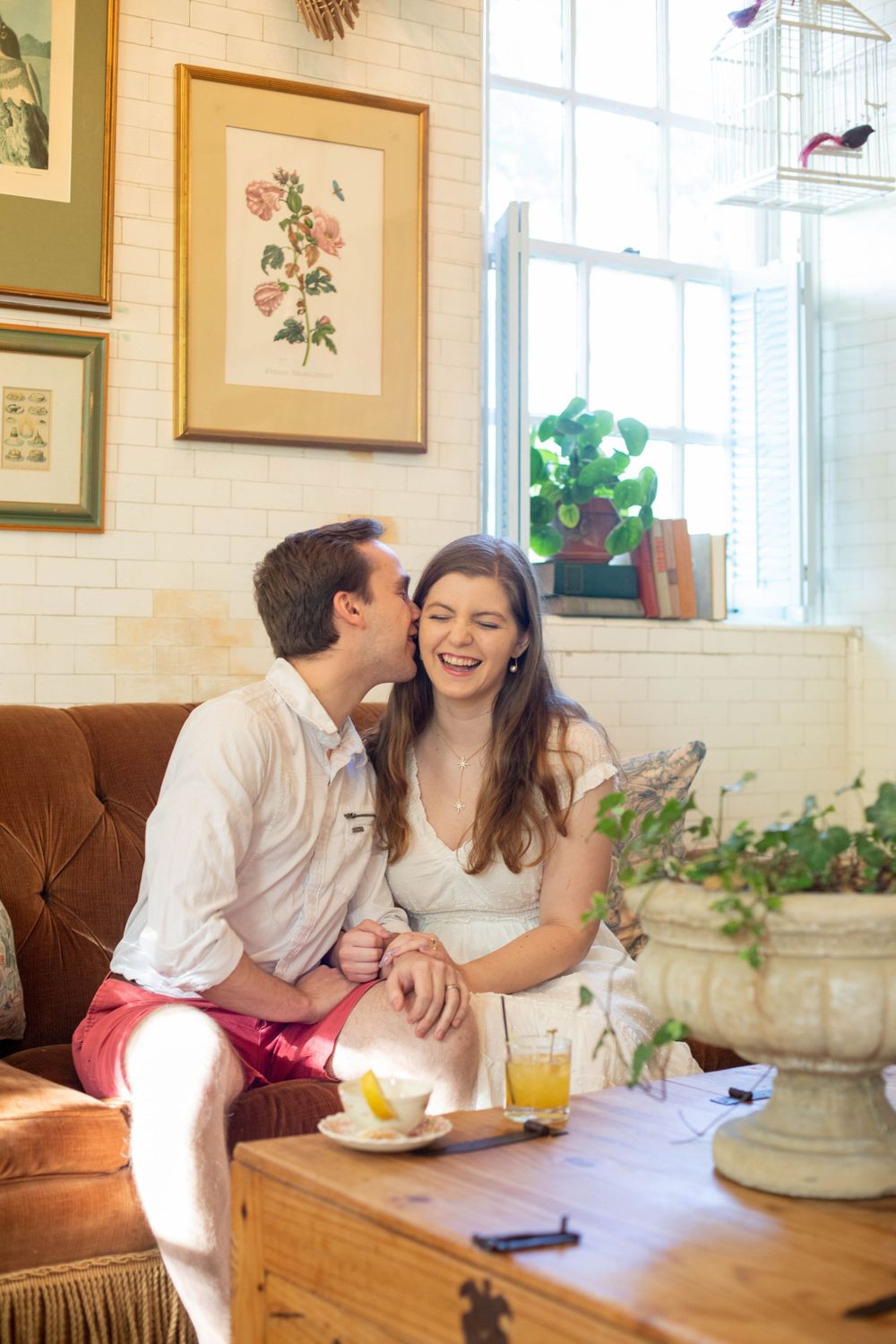A romantic couple shares intimate moments on a brown couch with botanical artwork on the walls behind them.