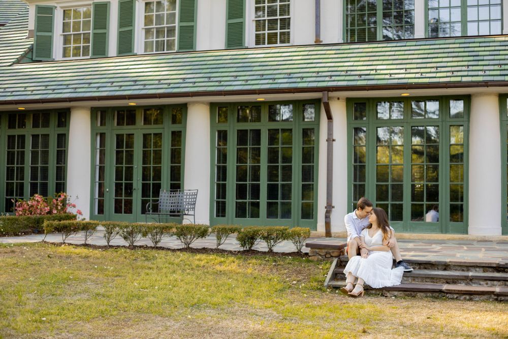 A couple poses romantically in front of a white building with green windows.