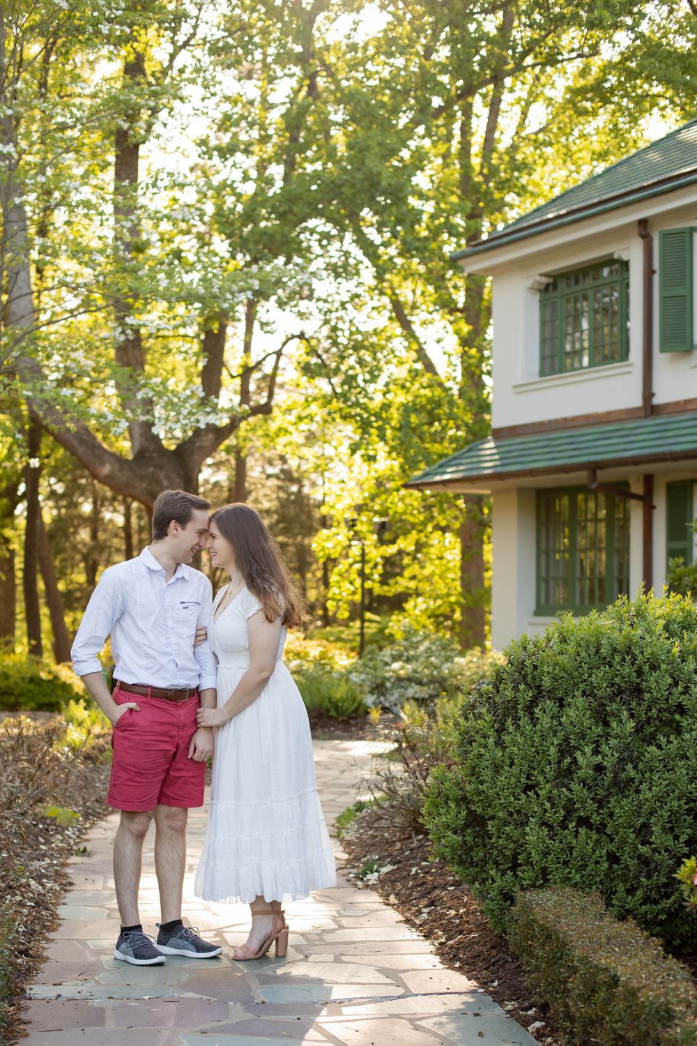 A couple in white and red poses along a garden path with beautiful sunlight filtering through trees.