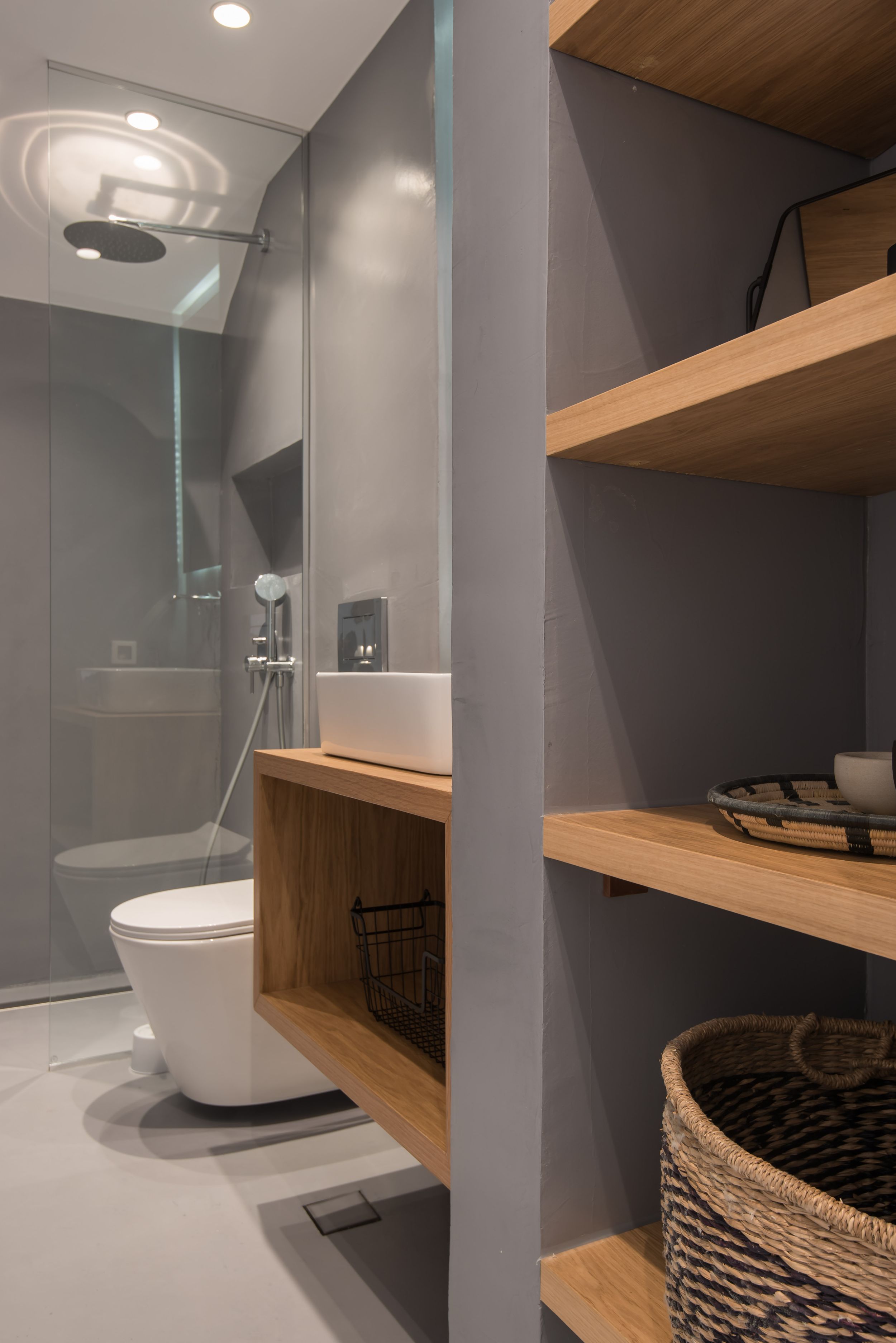Modern minimalist bathroom with gray walls, wooden shelving, and glass shower partition featuring clean lines and organized storage.