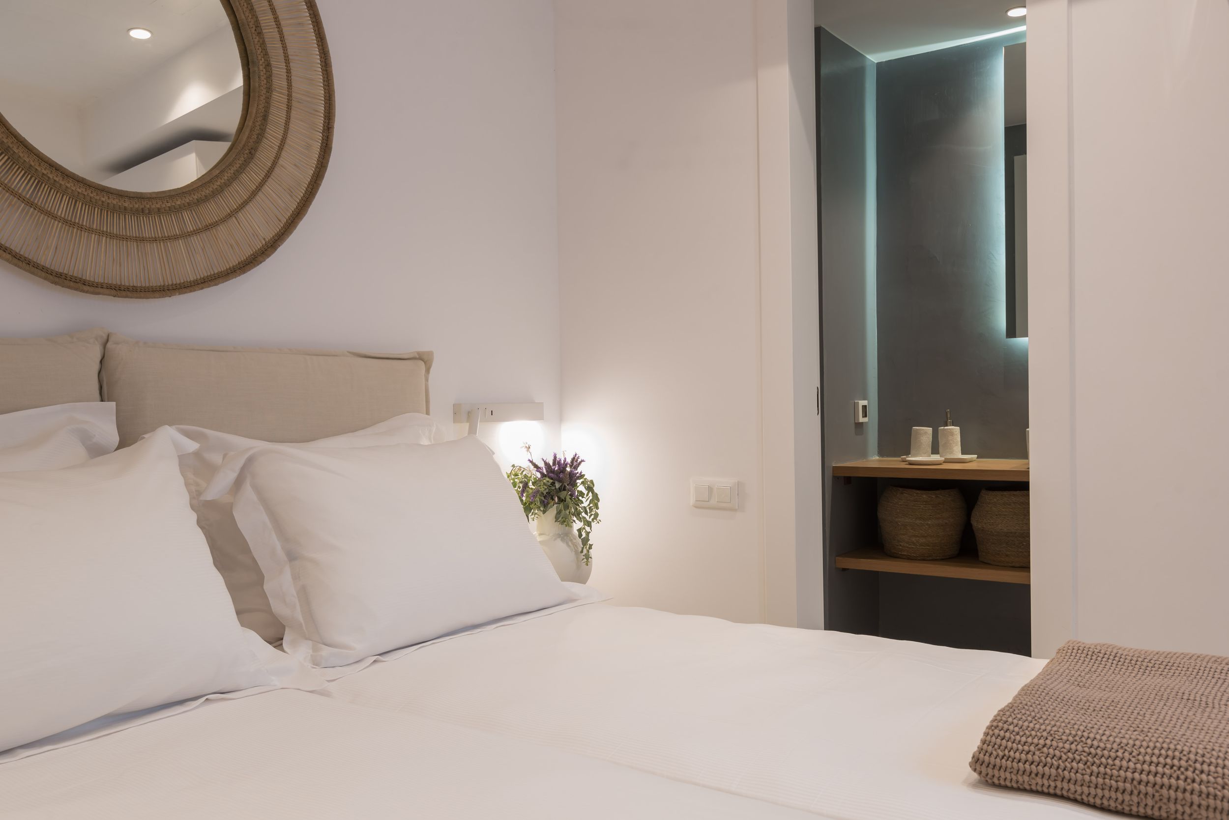 Modern hotel bedroom interior with white bedding, round mirror, and en-suite bathroom view illuminated by soft lighting.