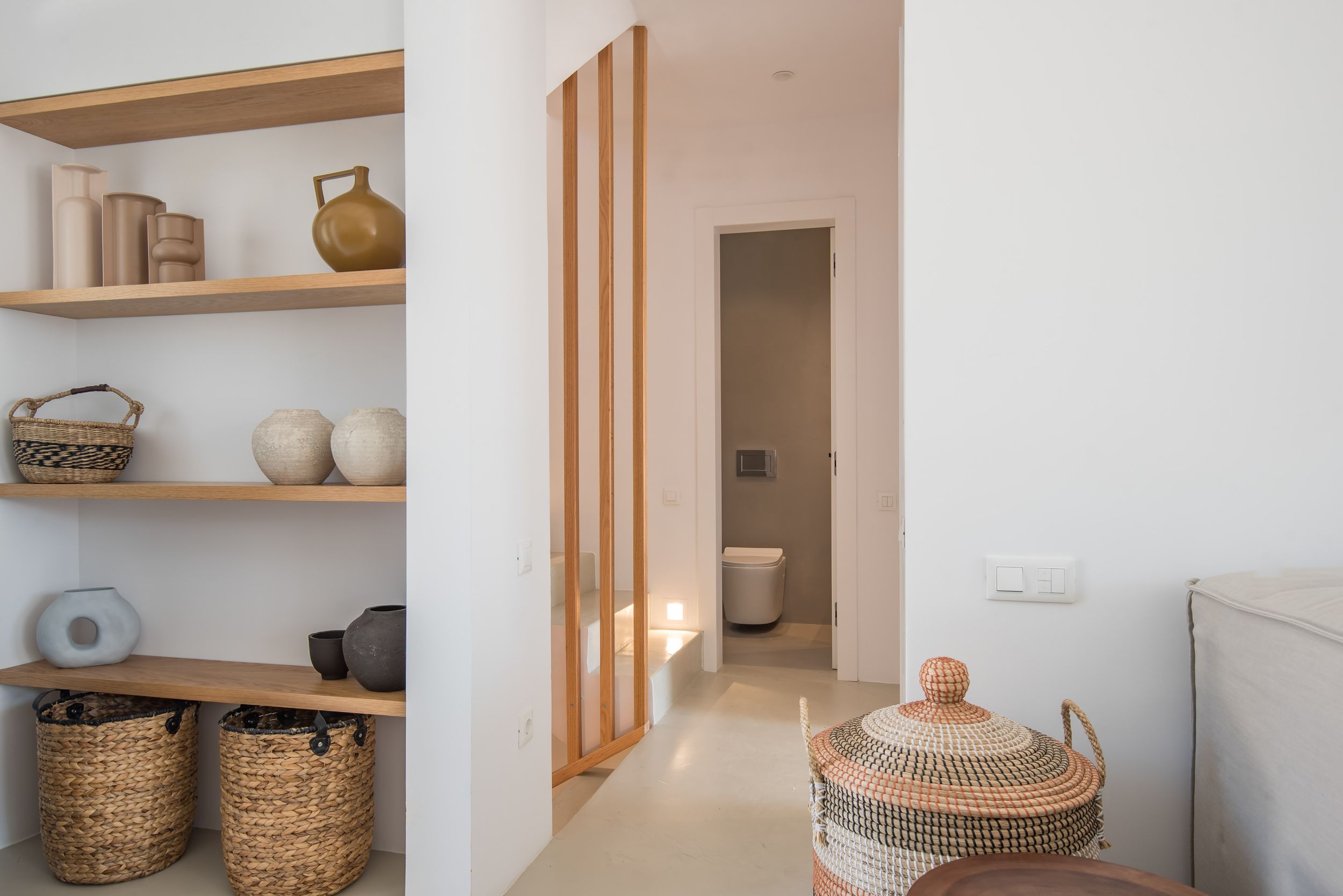 Modern minimalist interior with wooden slat wall detail, built-in shelving with woven baskets, and a hallway leading to bathroom.