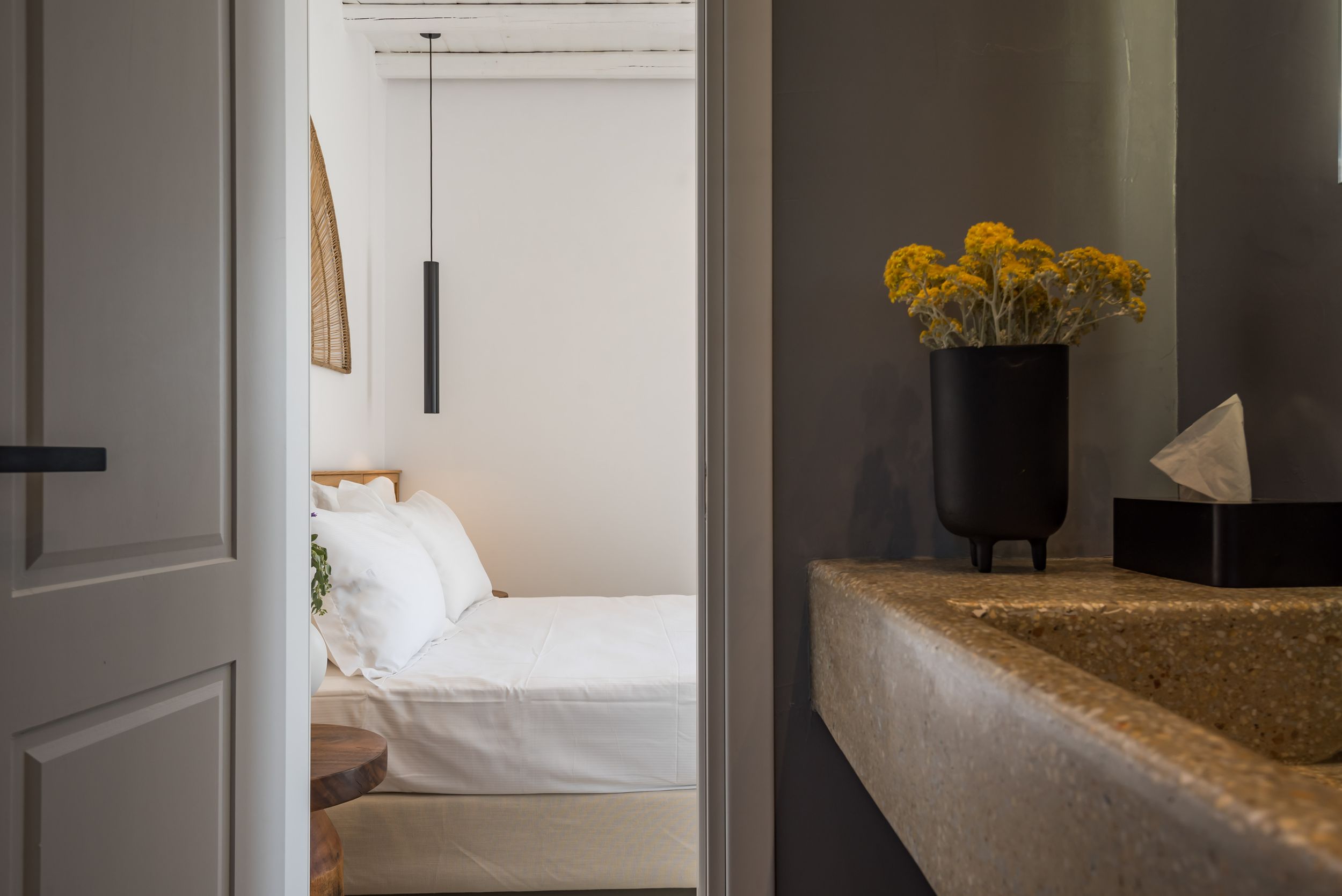 View through doorway into minimalist bedroom with white bedding and yellow flowers in black vase on granite counter.