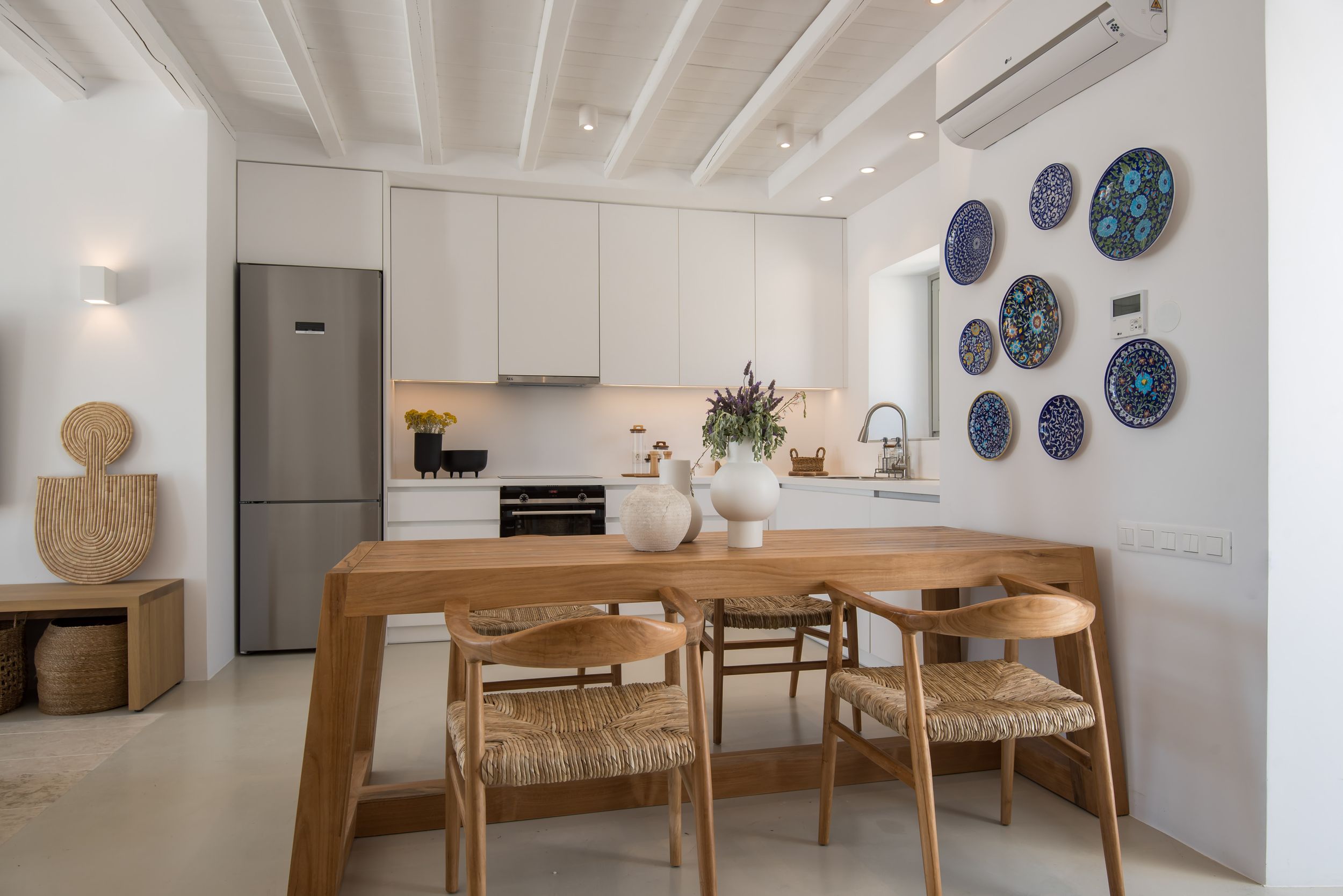 Modern Scandinavian kitchen with wooden dining table, woven chairs, white walls, and decorative blue plates mounted as wall art.