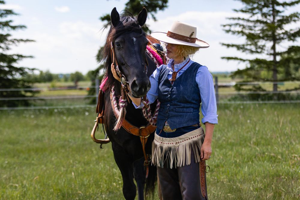 A horse stands with traditional western tack and gear in a grassy field with evergreen trees.