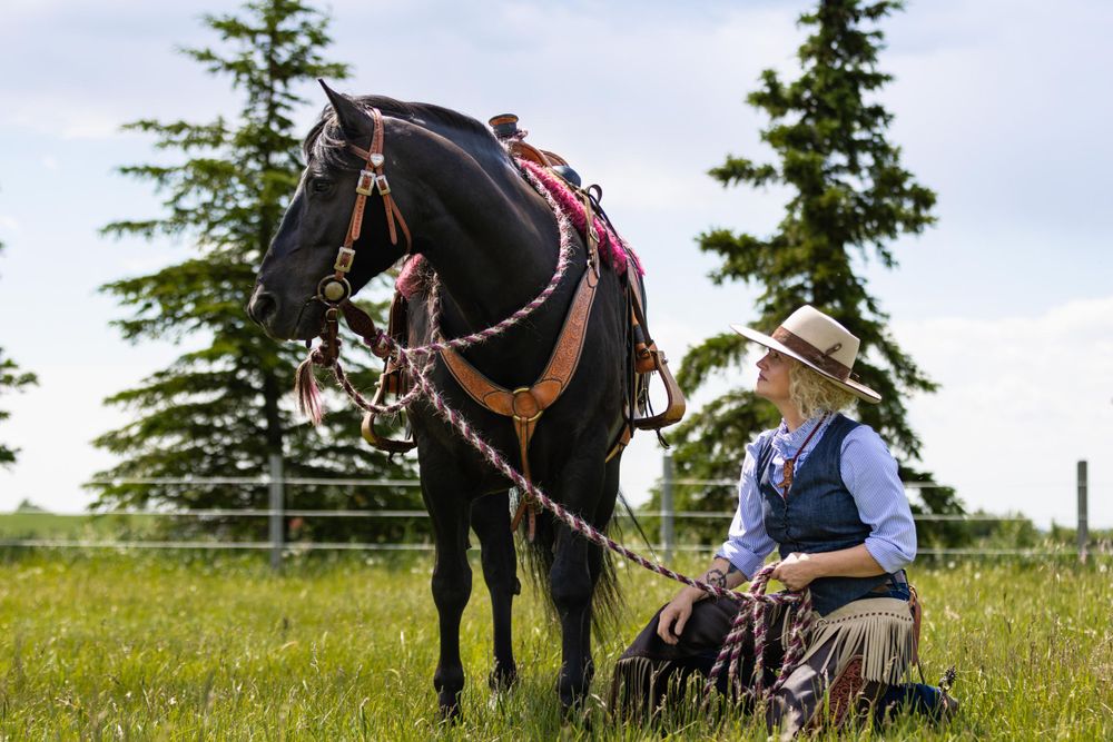 A dark horse with ornate leather tack and a cowgirl stand in a meadow with evergreen trees in the background.