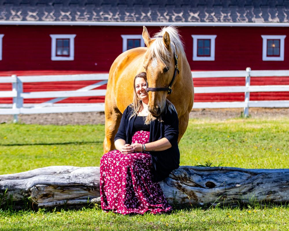 A palomino horse stands by a red barn with white trim on a sunny day.