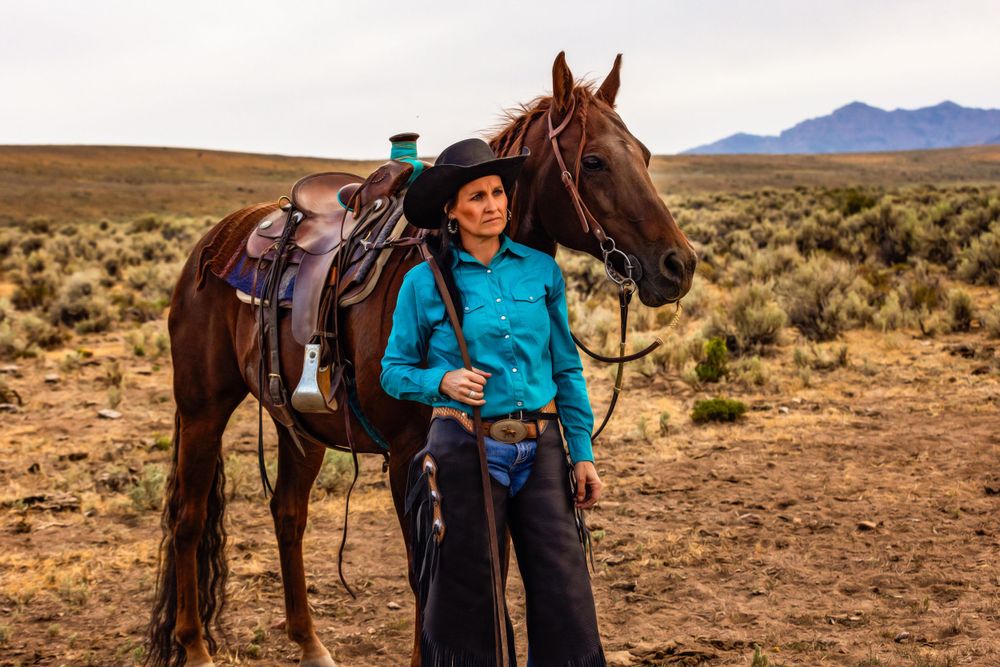 A brown horse with western tack and a cowgirl stand in the desert with mountains visible in the background.