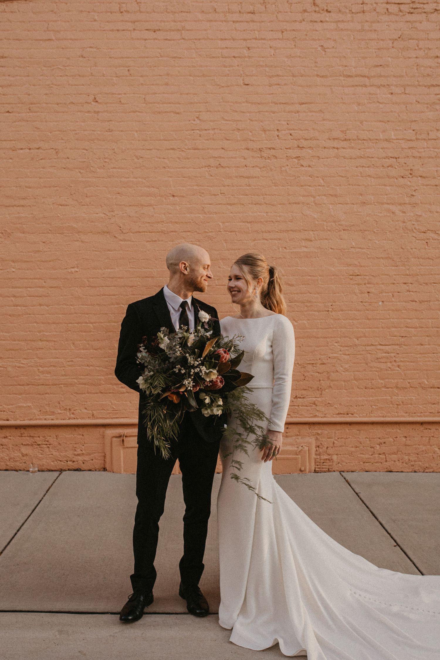 A bride and groom share a candid moment against a peachy brick wall, with the bride wearing a long-sleeved white gown.