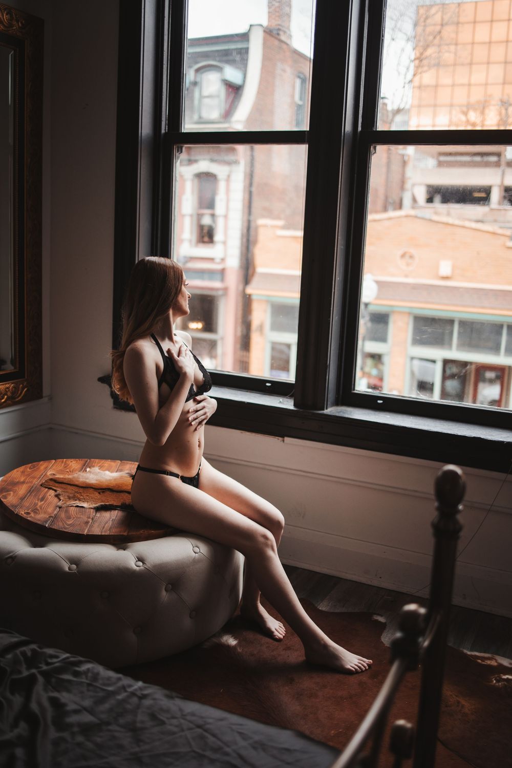 A moody boudoir photo of someone sitting on a bed by a window overlooking urban buildings.