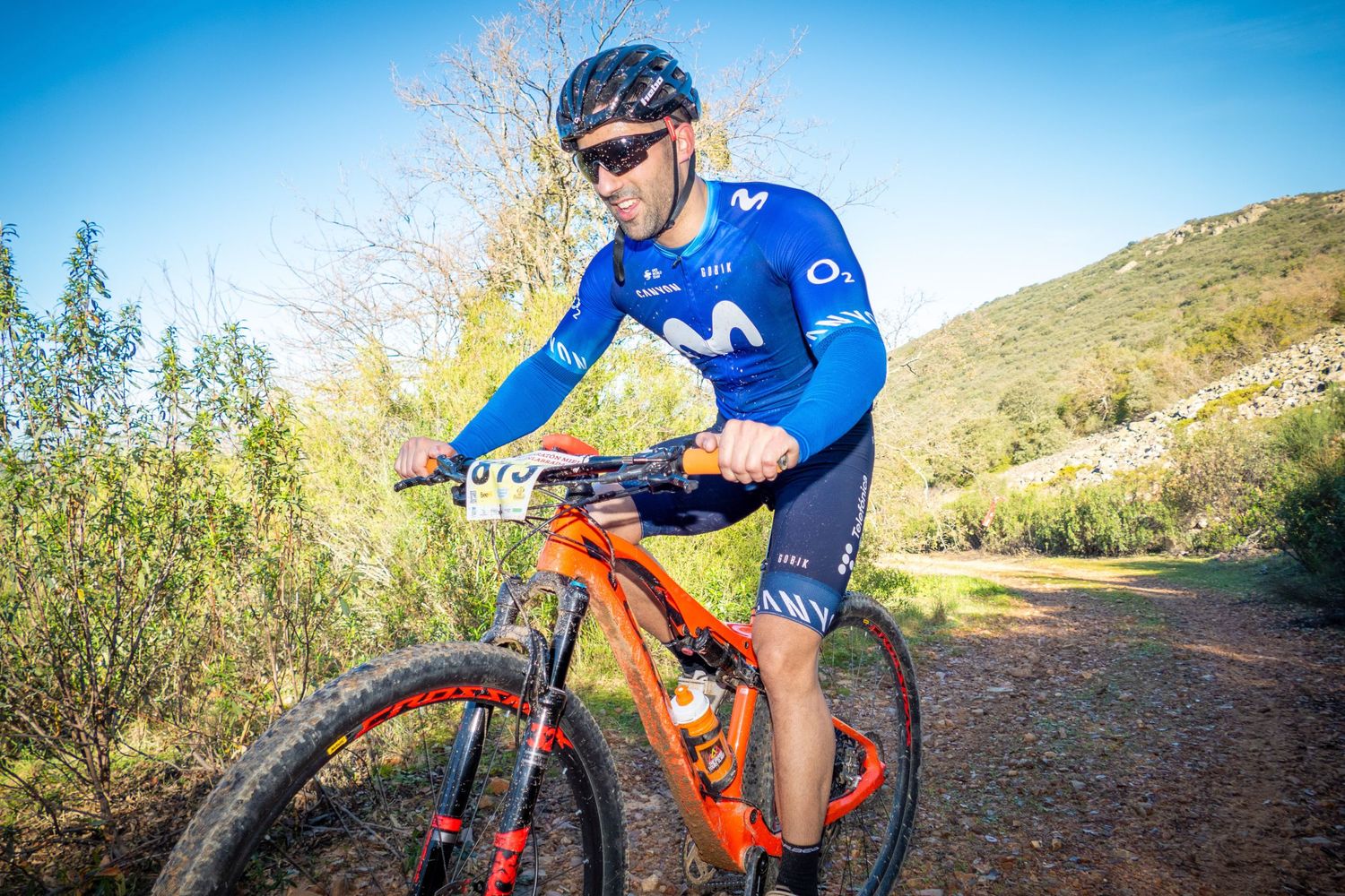 Cyclist in Movistar team kit rides orange mountain bike on rugged trail with blue sky backdrop.