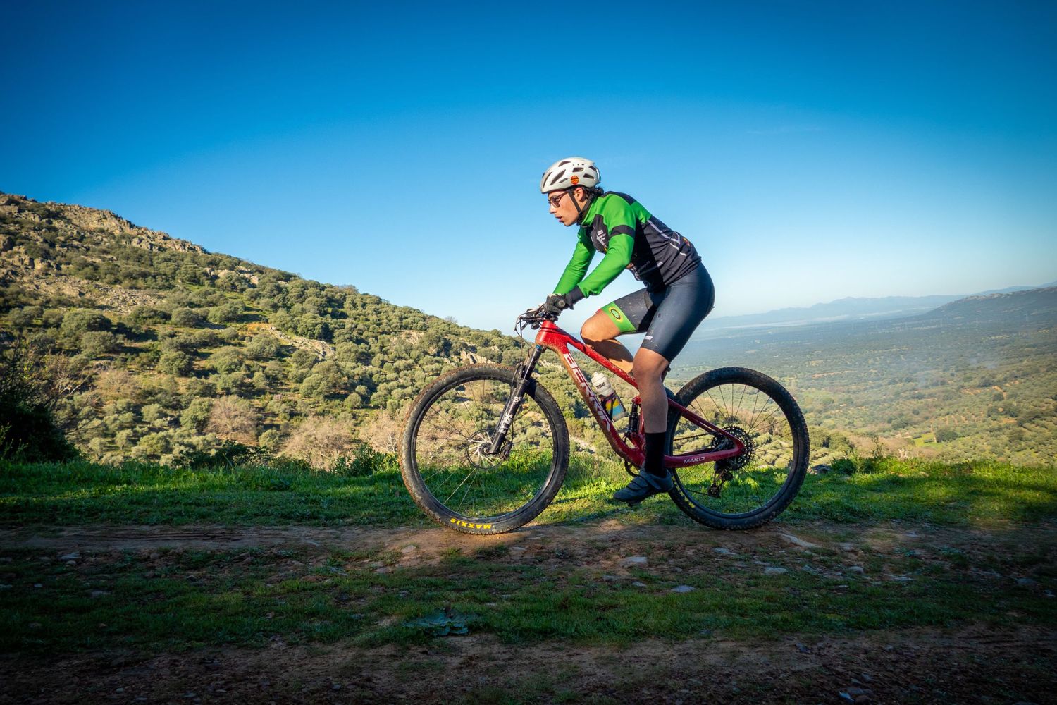 Mountain biker in green jersey rides red bike on dirt trail with scenic green hills in background.