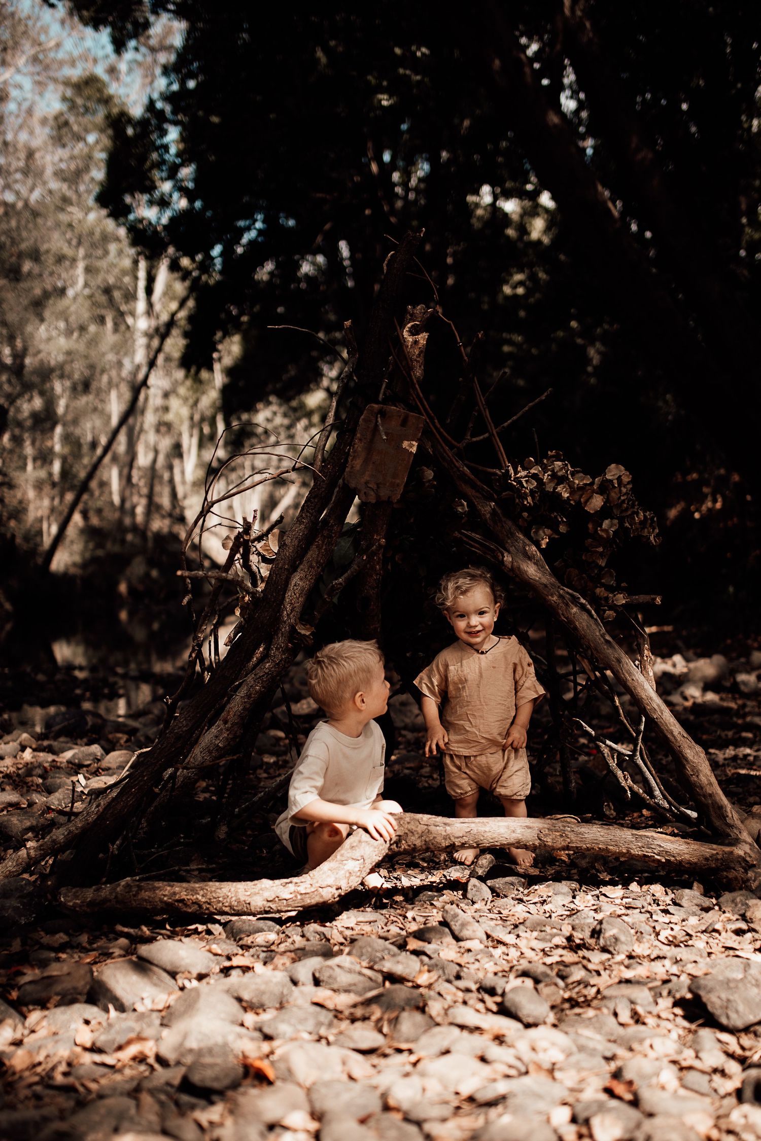 Two small children sit beneath a rustic wooden fort made of sticks and branches in a woodland setting.