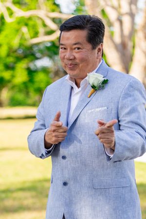 Person in light gray suit with boutonniere points cheerfully at camera during outdoor event. Merritt Island, Florida