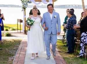 Newlyweds walk down aisle under purple-draped arch with water view in background. Merritt Island, Florida