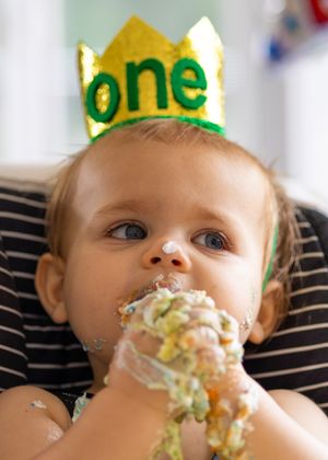 A baby wearing a green 'one' crown enjoys their first birthday cake with messy frosting. West Palm Beach, Florida