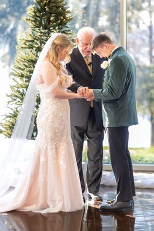 Bride in a blush lace gown exchanges rings with groom in green jacket during indoor winter wedding ceremony. Lake Mary, Florida