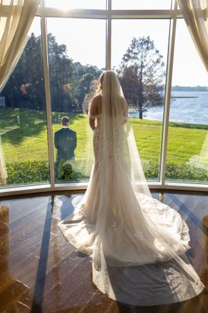 Bride in flowing white dress and veil gazes out large window overlooking waterfront landscape. Lake Mary, Florida