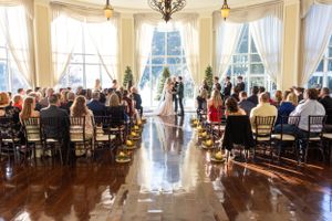 Elegant wedding ceremony venue with tall windows, white curtains and rows of gold chairs filled with guests. Lake Mary, Florida