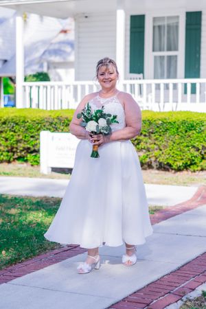 A bride in a flowing white tea-length dress holds a bouquet of white flowers on a porch with green shutters. Merritt Island, Florida